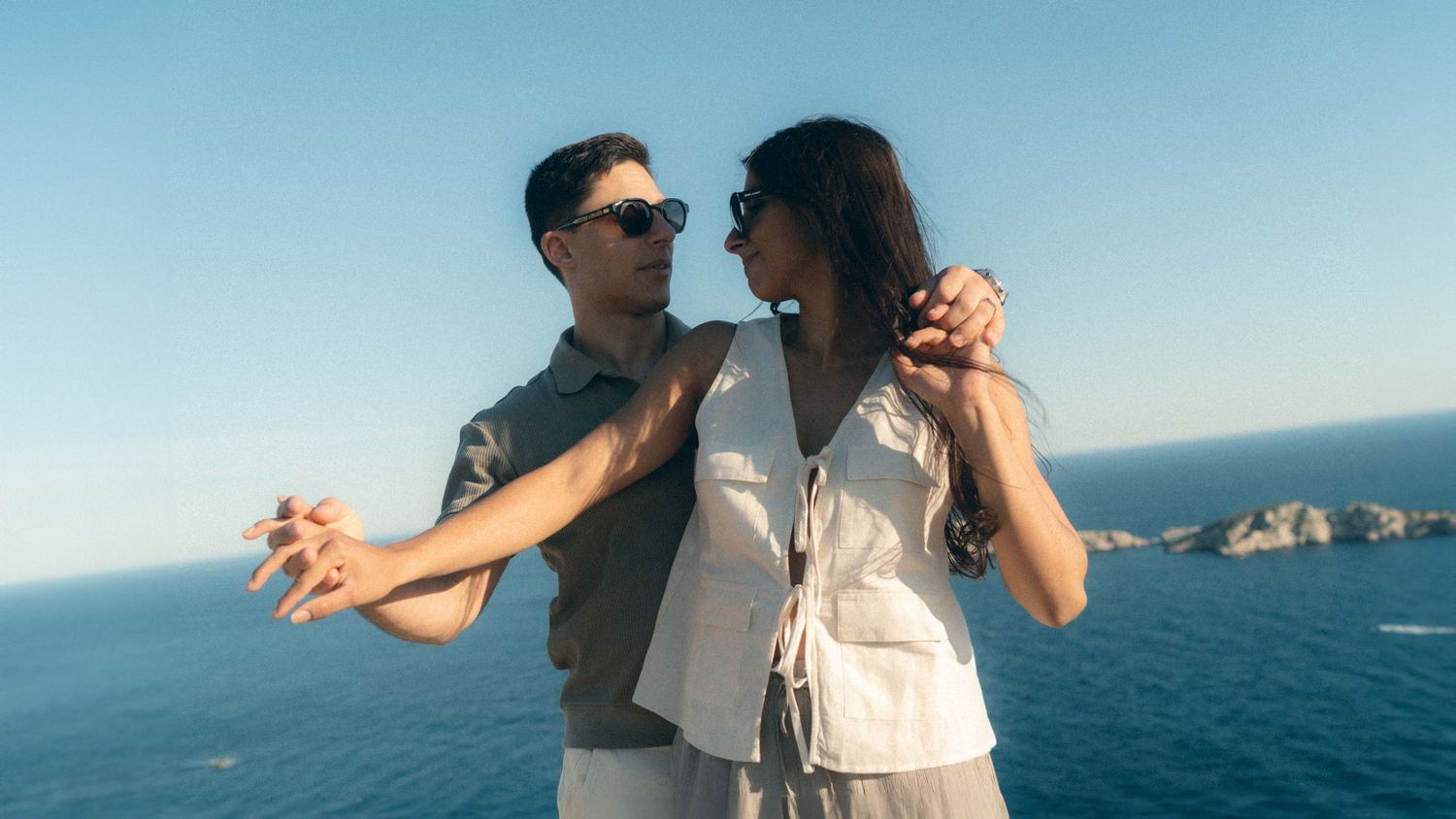 A couple shares a romantic moment while dancing together on a coastal cliff overlooking the blue ocean.