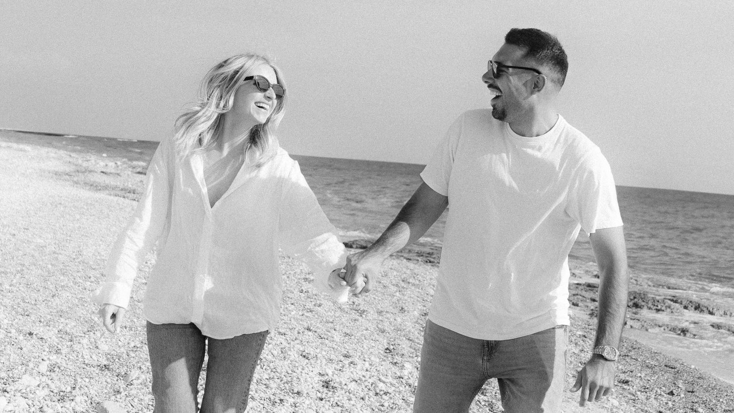 A couple walks hand in hand along a sandy beach shoreline on a sunny day.