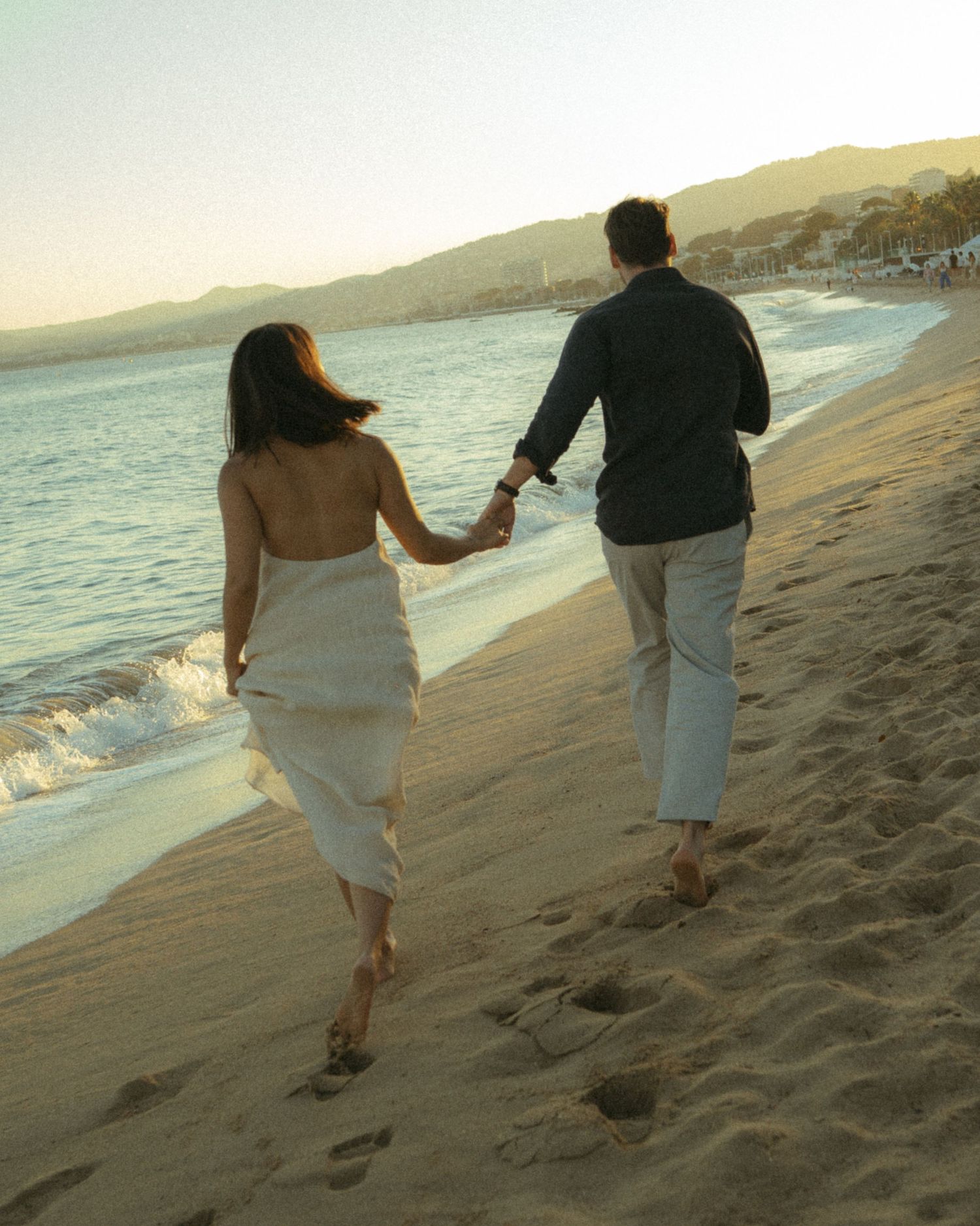Couple walking together along a scenic beach at sunset holding wine glasses and leaving footprints in the sand.