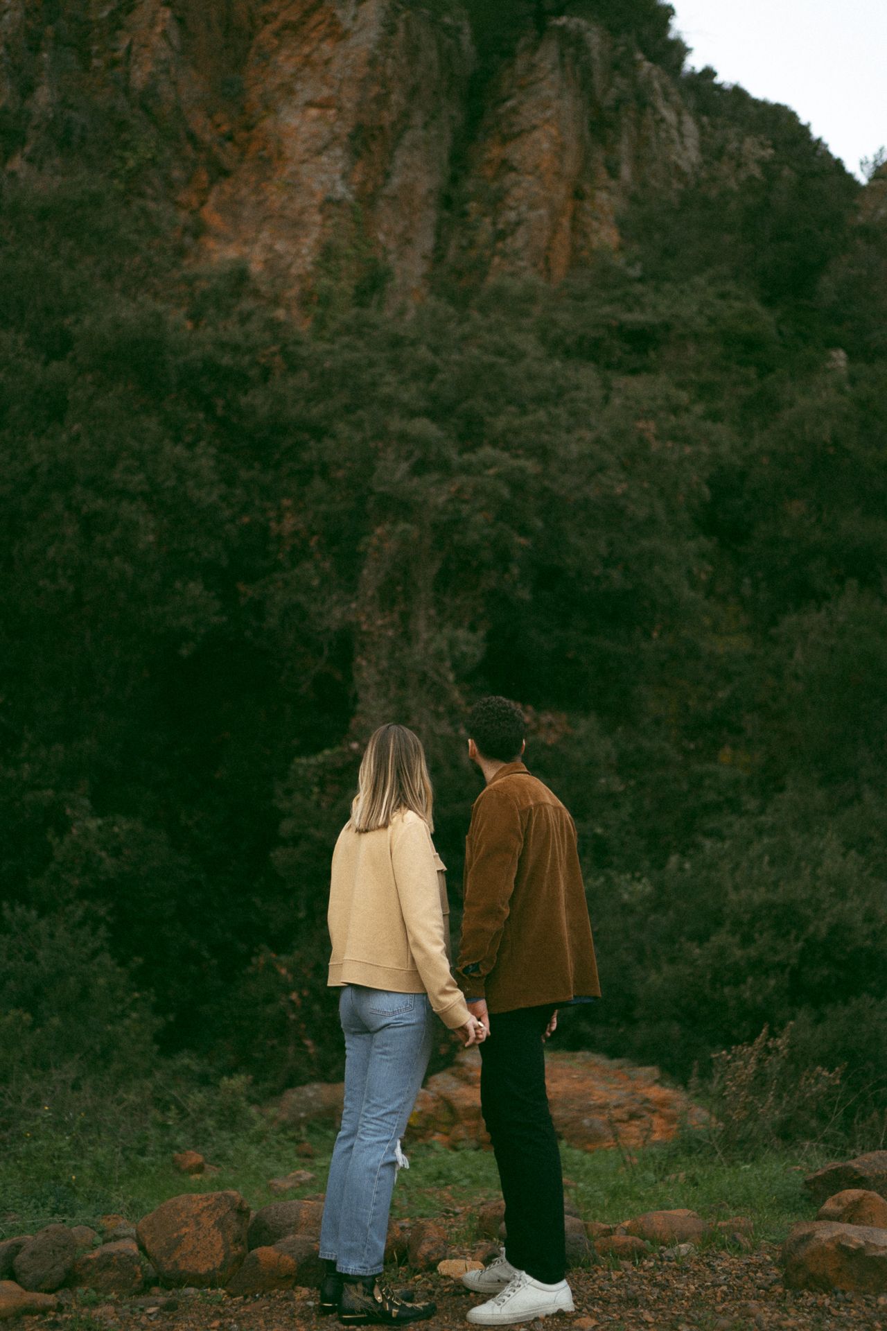 Two people stand at the edge of a rocky cliff during golden hour against a dark mountainous backdrop.
