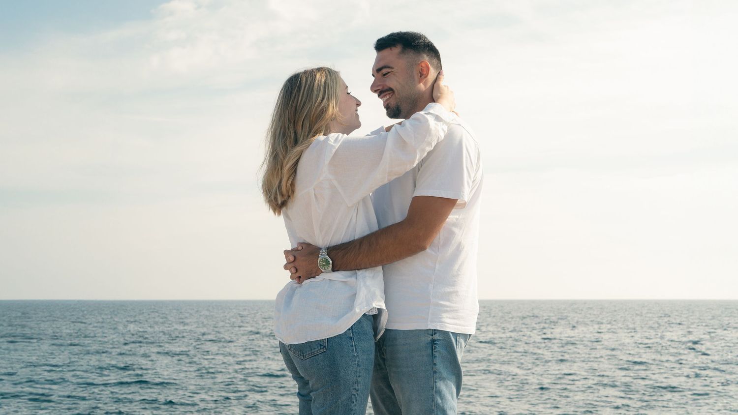 Romantic couple in white outfits embracing on beach at sunset with ocean waves in background.