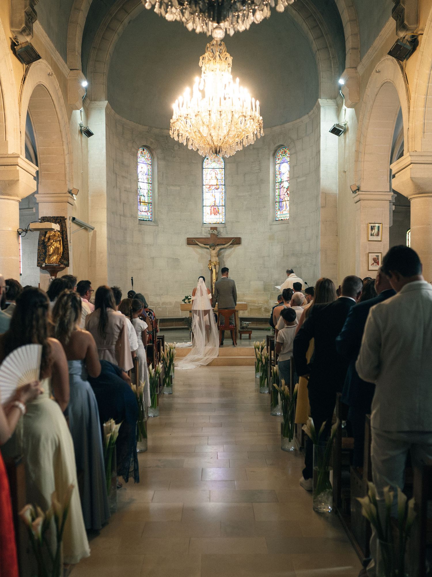 Elegant church wedding ceremony with guests seated in pews, floral decorations, crystal chandelier, and couple at the altar.
