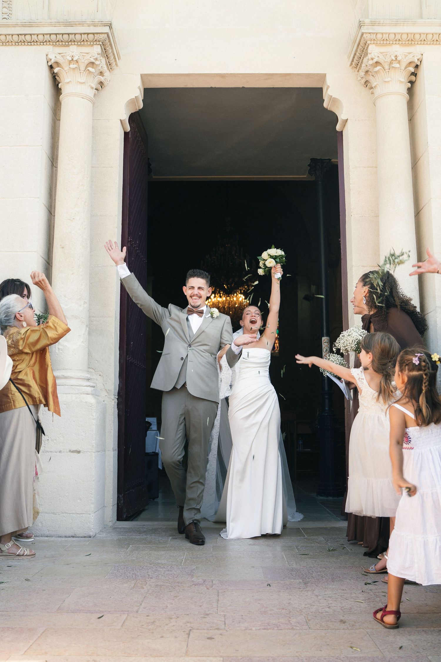 Joyful newlyweds exit church doors, bride holding bouquet aloft, groom waving, greeted by cheering guests and flower girl.