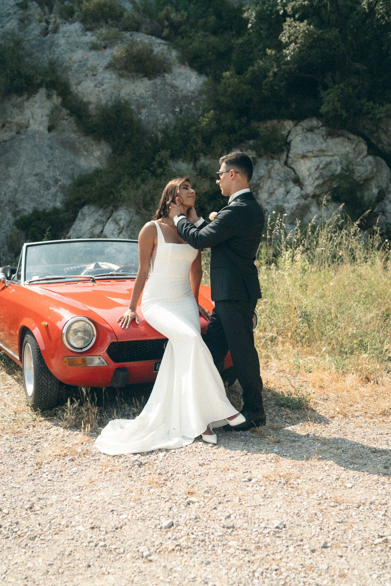 A bride and groom pose for romantic wedding photos next to a vintage red sports car on a gravel road.