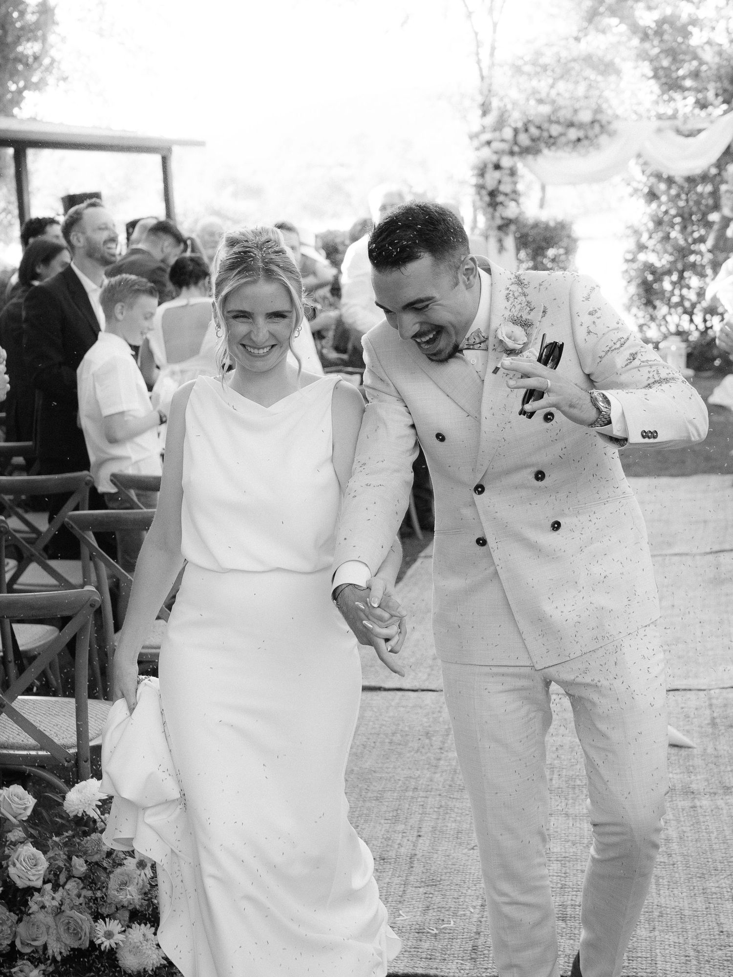 Black and white photo of a joyful bride and groom holding hands, laughing as they walk away from their wedding ceremony.