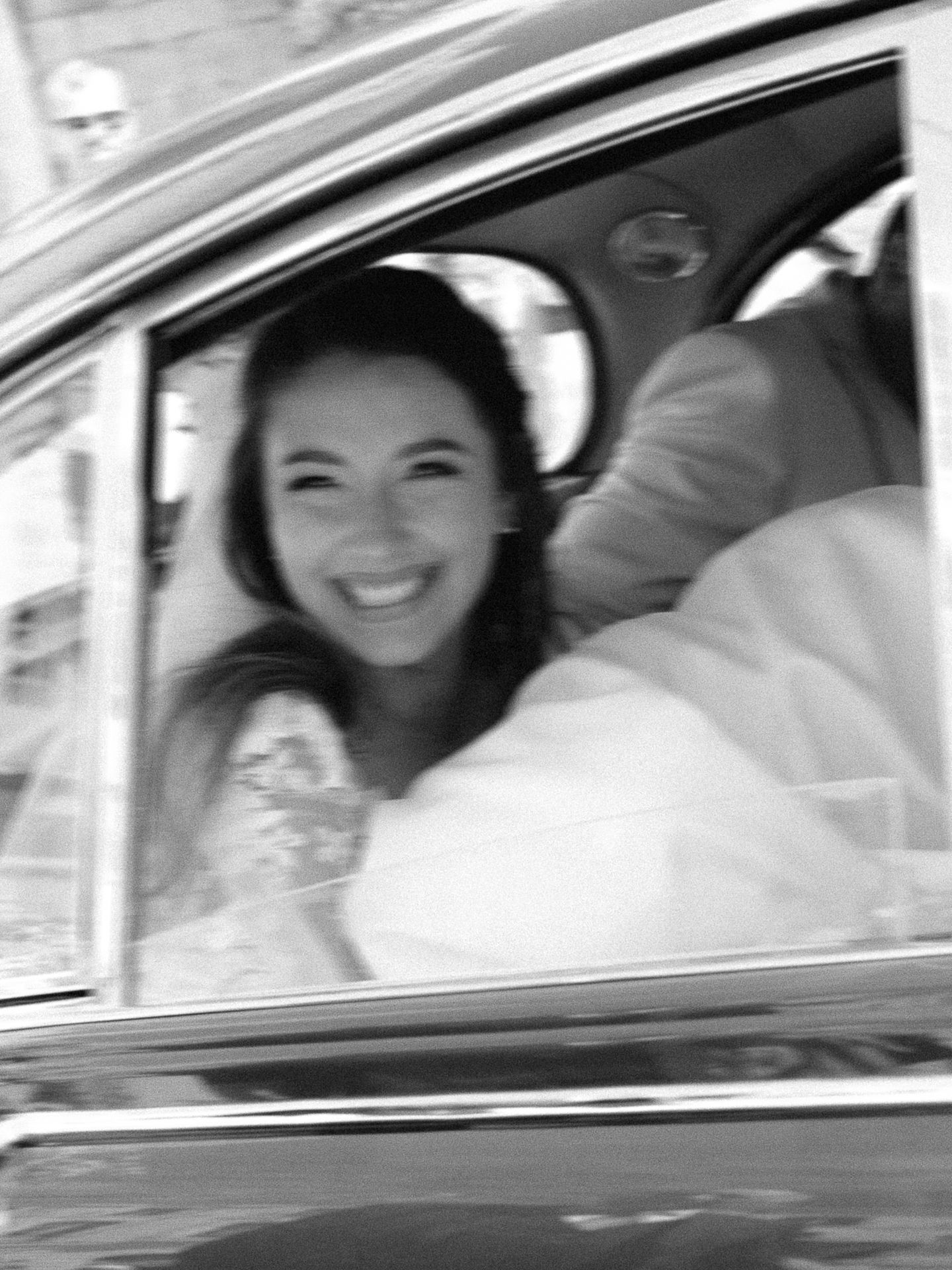 Smiling bride in wedding dress sitting in a classic car, captured in black and white photography.