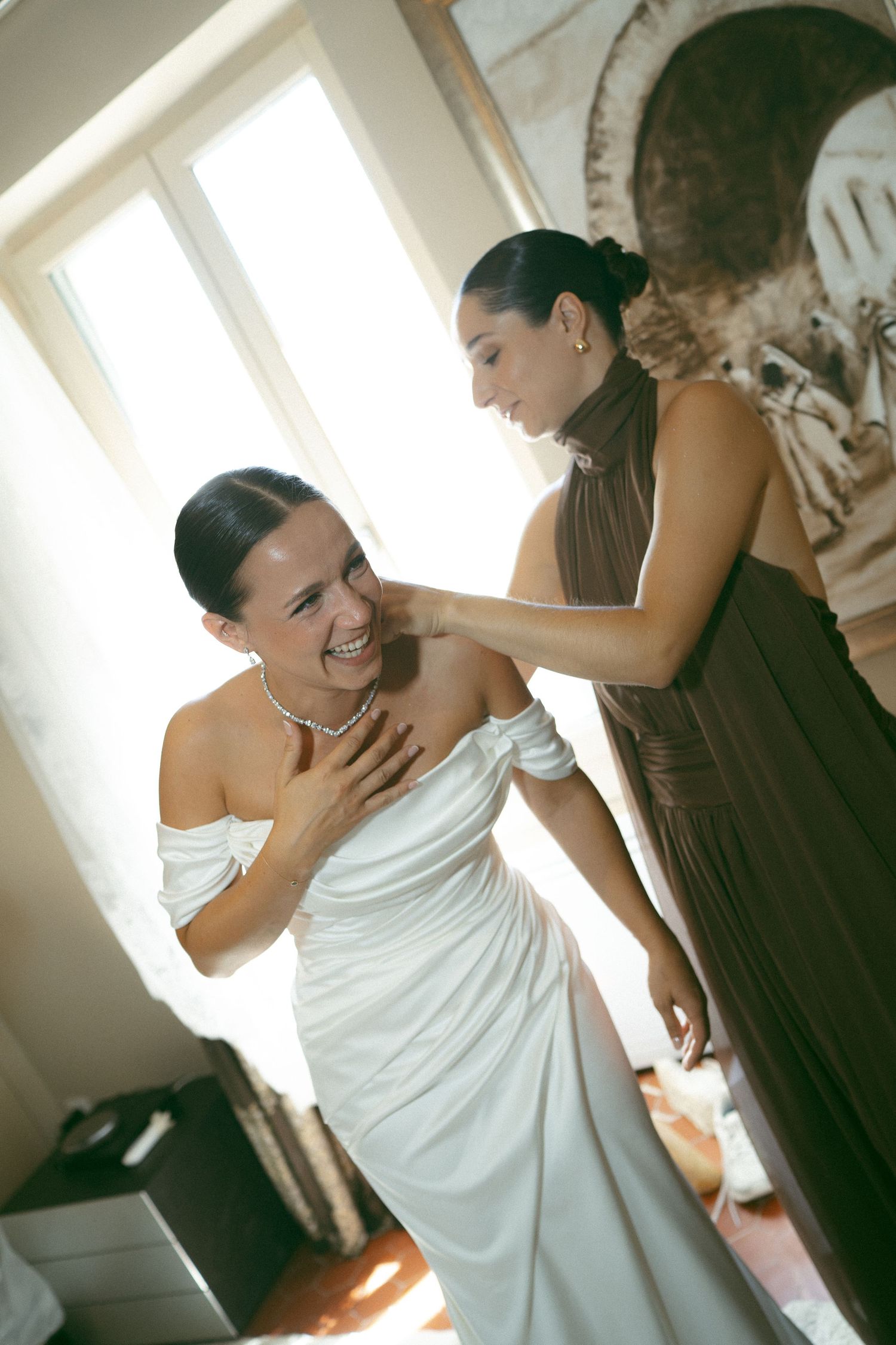 Bride in white off-shoulder gown laughing as bridesmaid in brown dress helps her prepare for wedding ceremony.