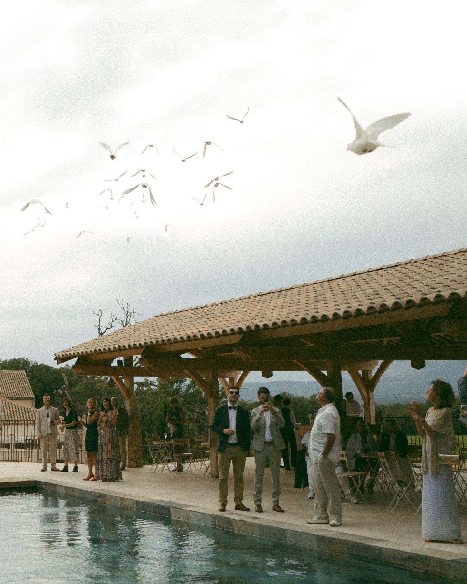 Guests gather under a covered outdoor patio area overlooking a swimming pool with mountain views in the background.
