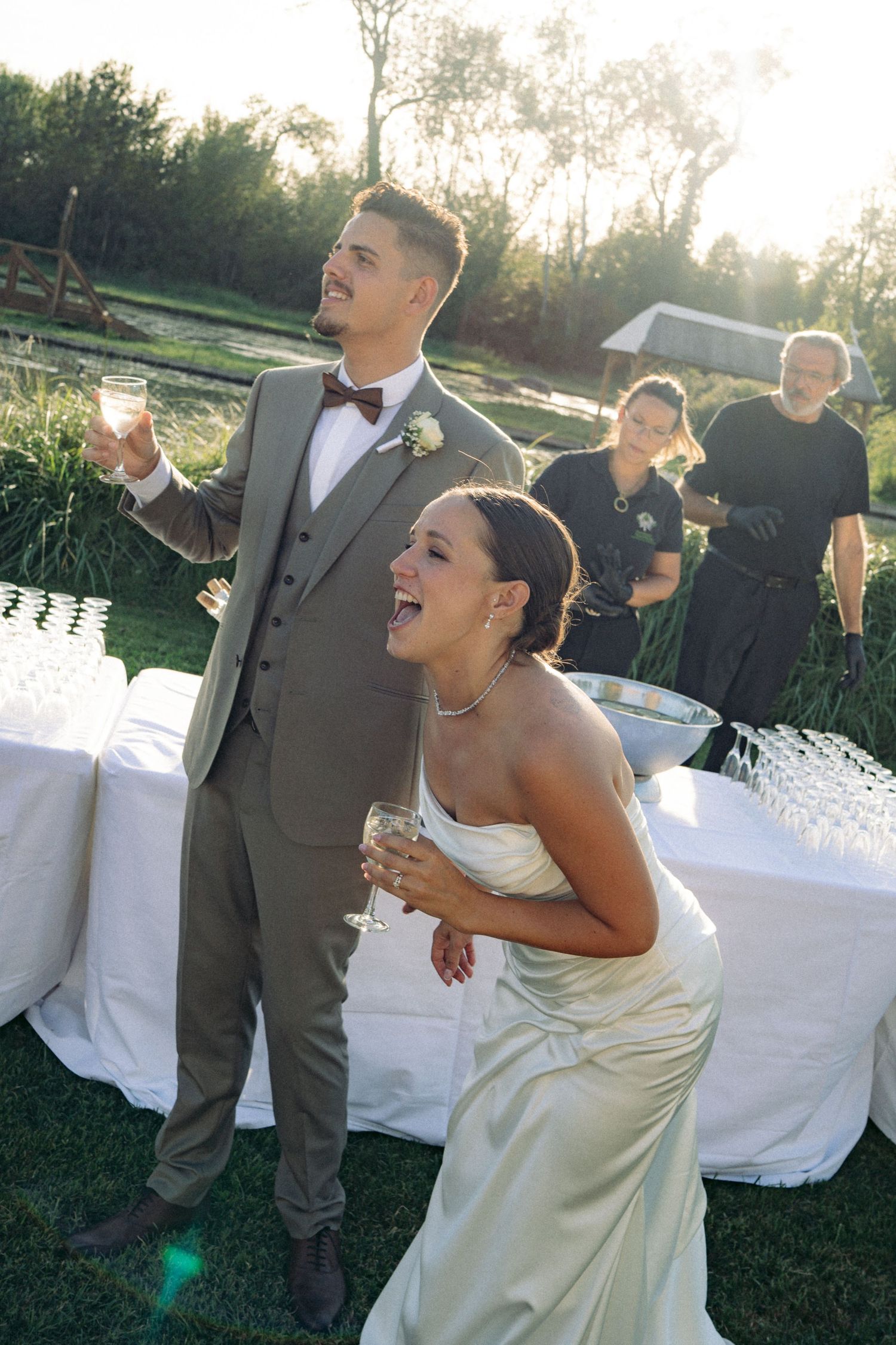 Bride and groom laughing and raising champagne glasses outdoors at golden hour wedding reception.