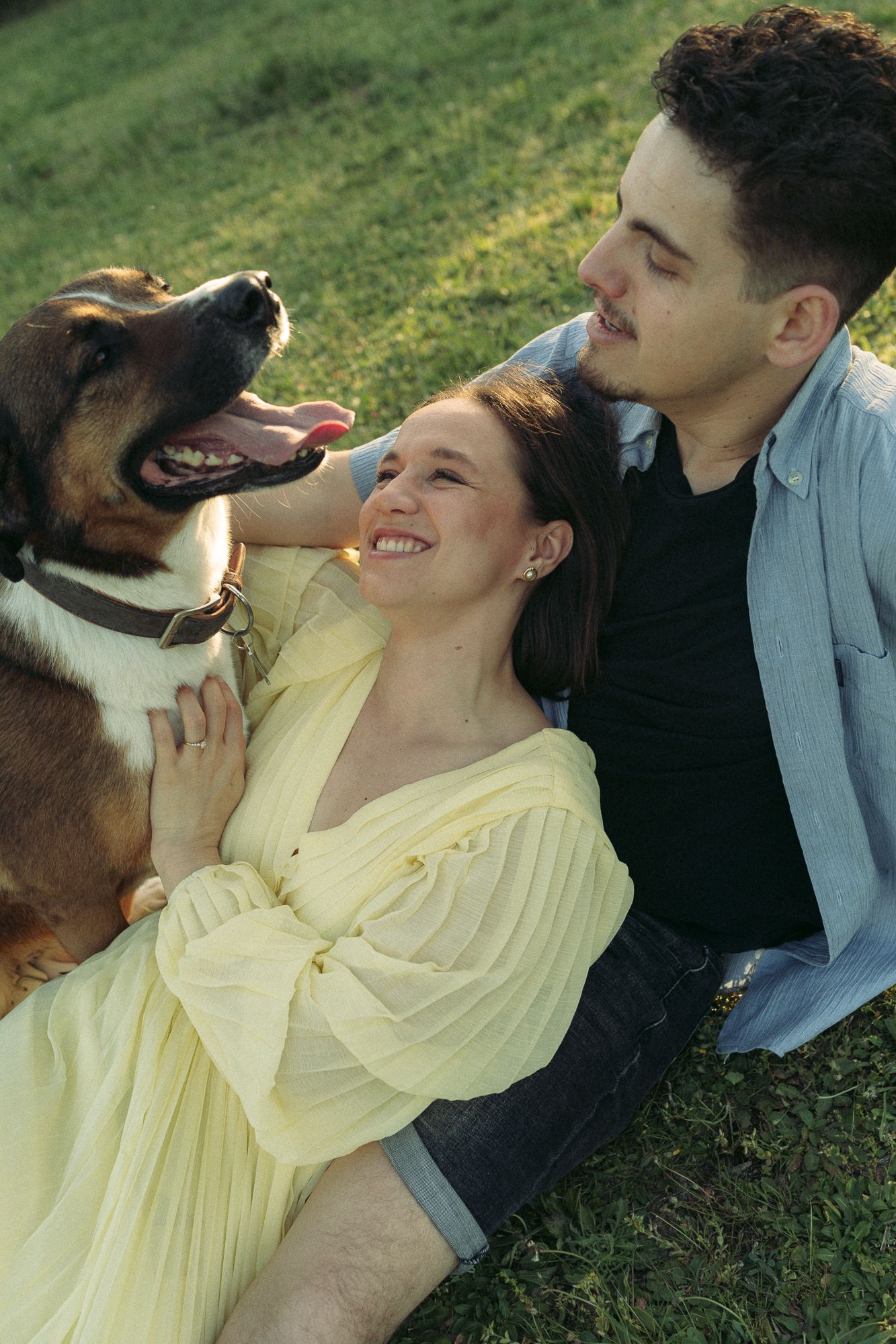 A smiling couple relaxes on green grass with their happy dog, the woman in a yellow dress leaning back on the man.