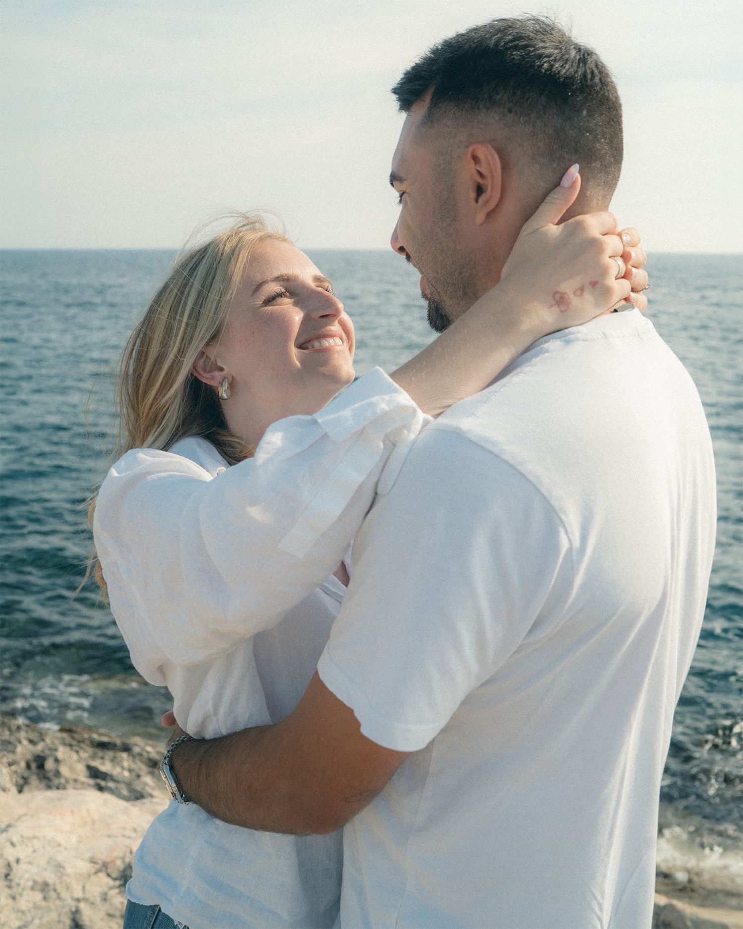 A couple in white clothing embraces while standing near the ocean on a sunny day.