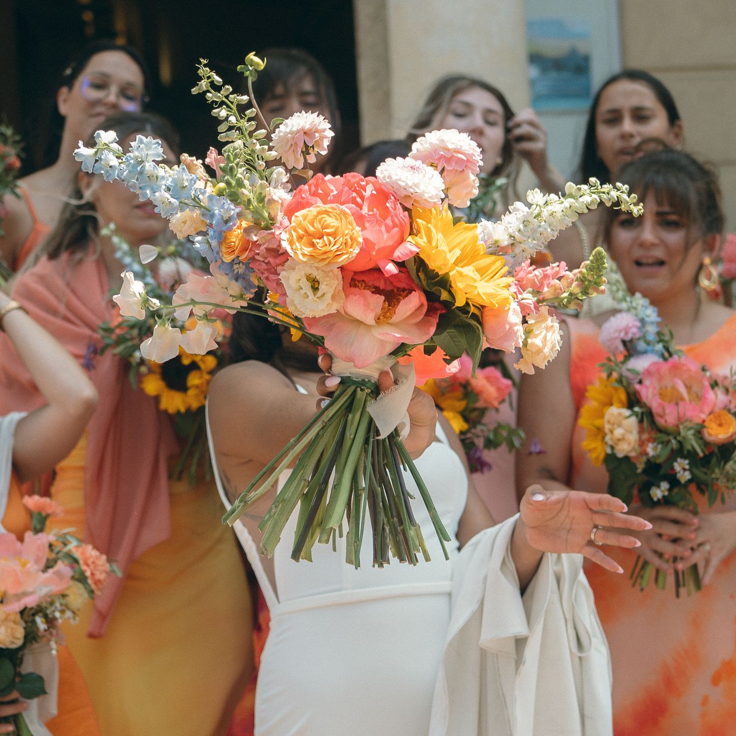 Wedding party members holding colorful coral and peach floral bouquets against a soft blurred background.
