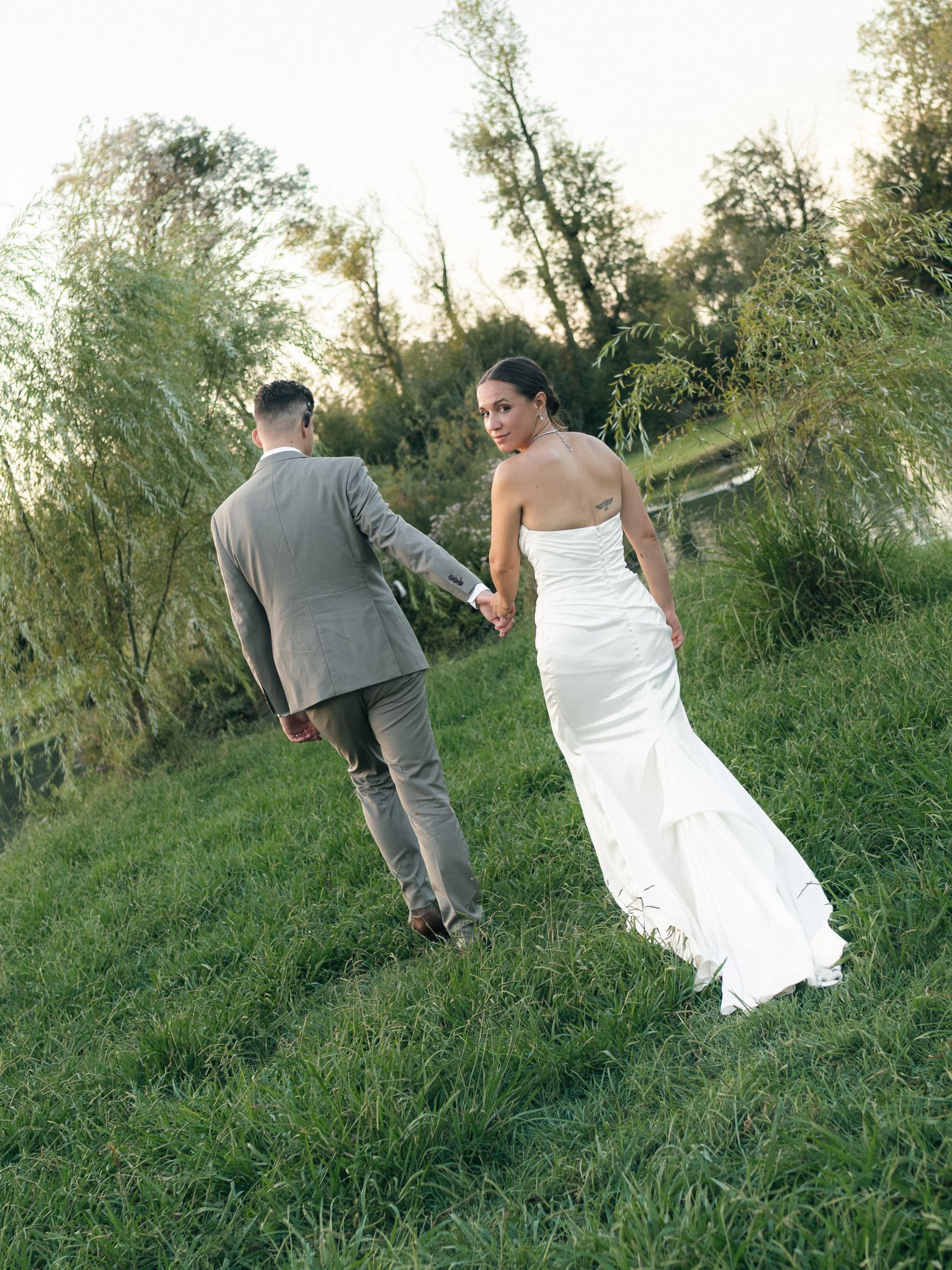 A bride in a white gown and groom in a tan suit walk hand-in-hand across a lush green lawn near a pond.