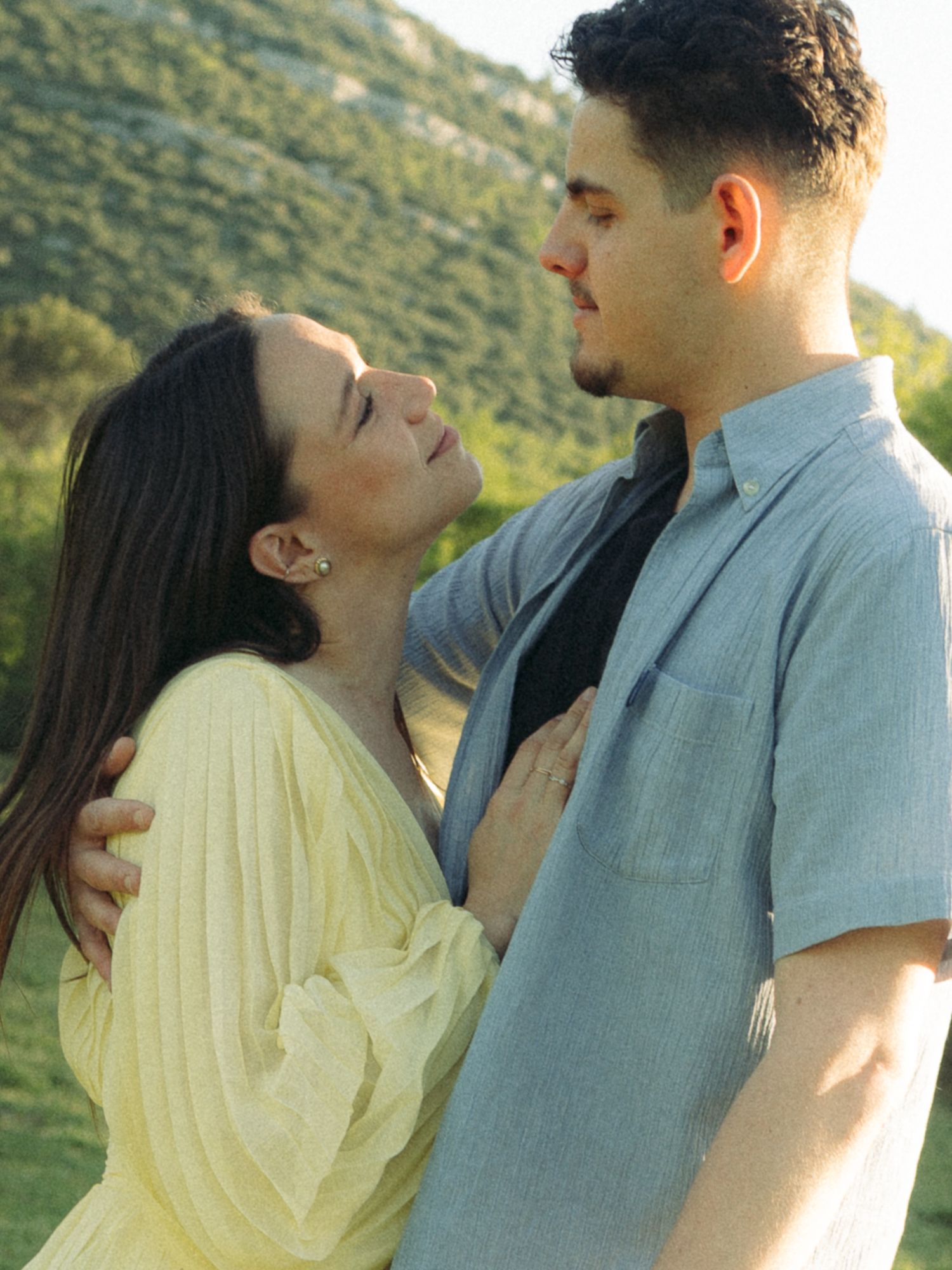 A couple shares a romantic moment outdoors, with green hills in the background, as they gaze at each other closely.