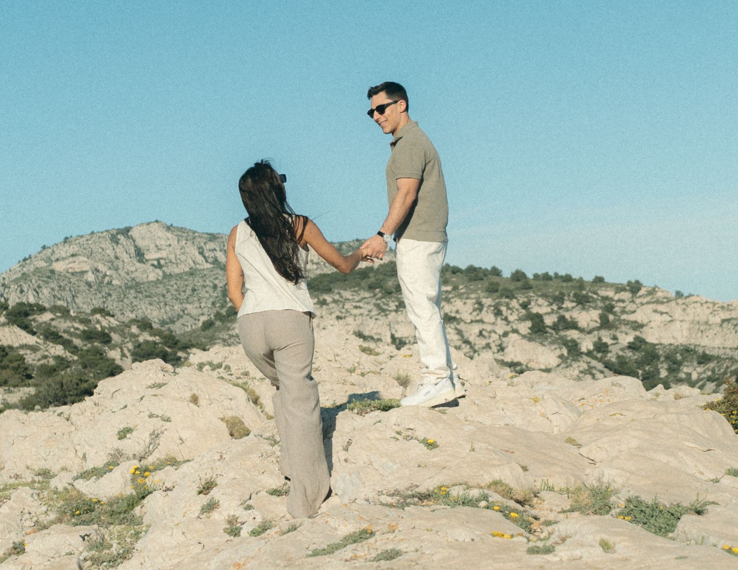 Two people hold hands while hiking on a rocky desert mountain trail against a bright blue sky.
