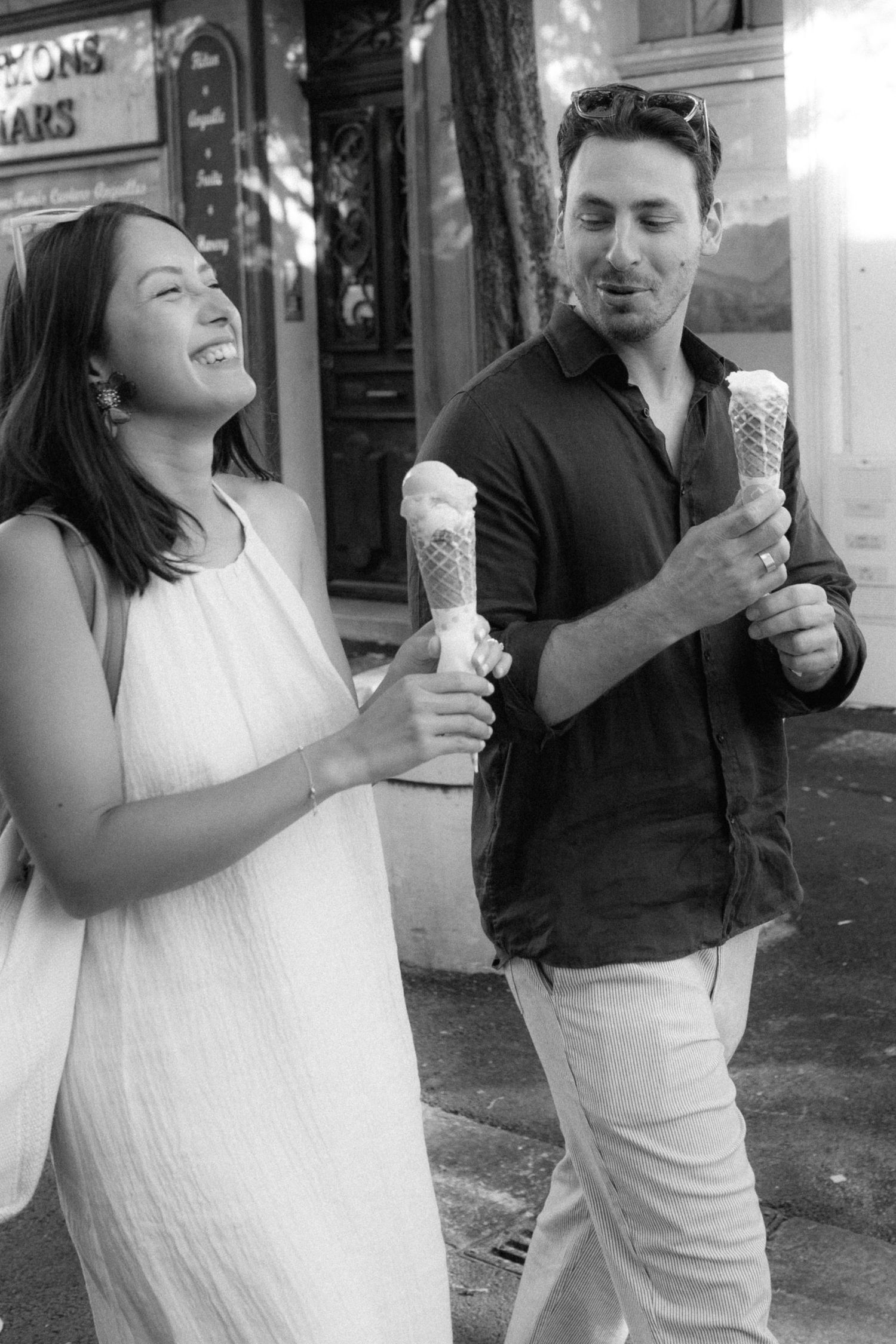 A couple shares a joyful moment while eating ice cream cones together on a city street.