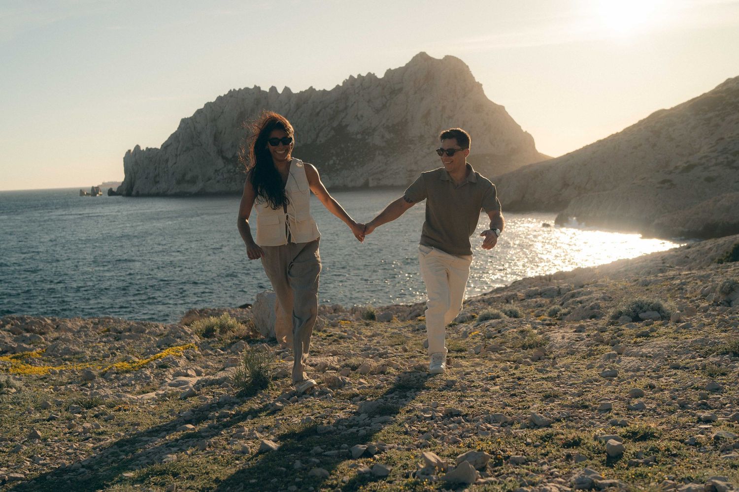A couple walks along a rocky coastal beach at sunset with mountains in the background.