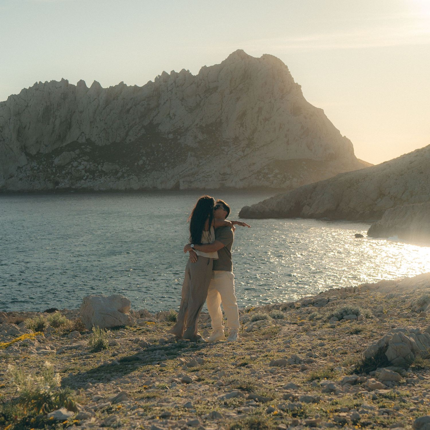 Two figures stand on a rocky coastline at sunset as golden light illuminates the mountainous landscape and calm waters.