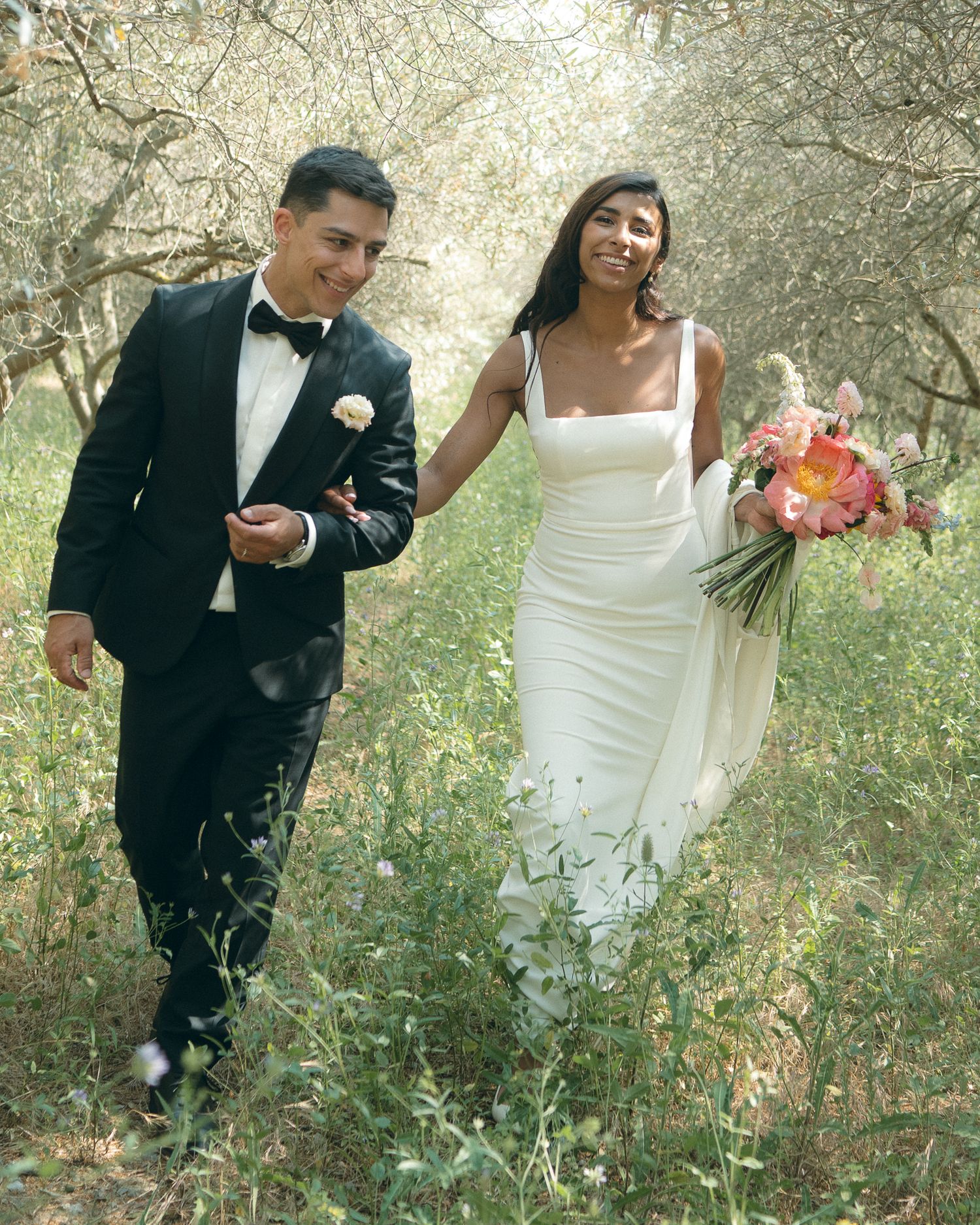 A couple in wedding attire shares joyful moments while walking through a sunlit garden with trees and greenery.