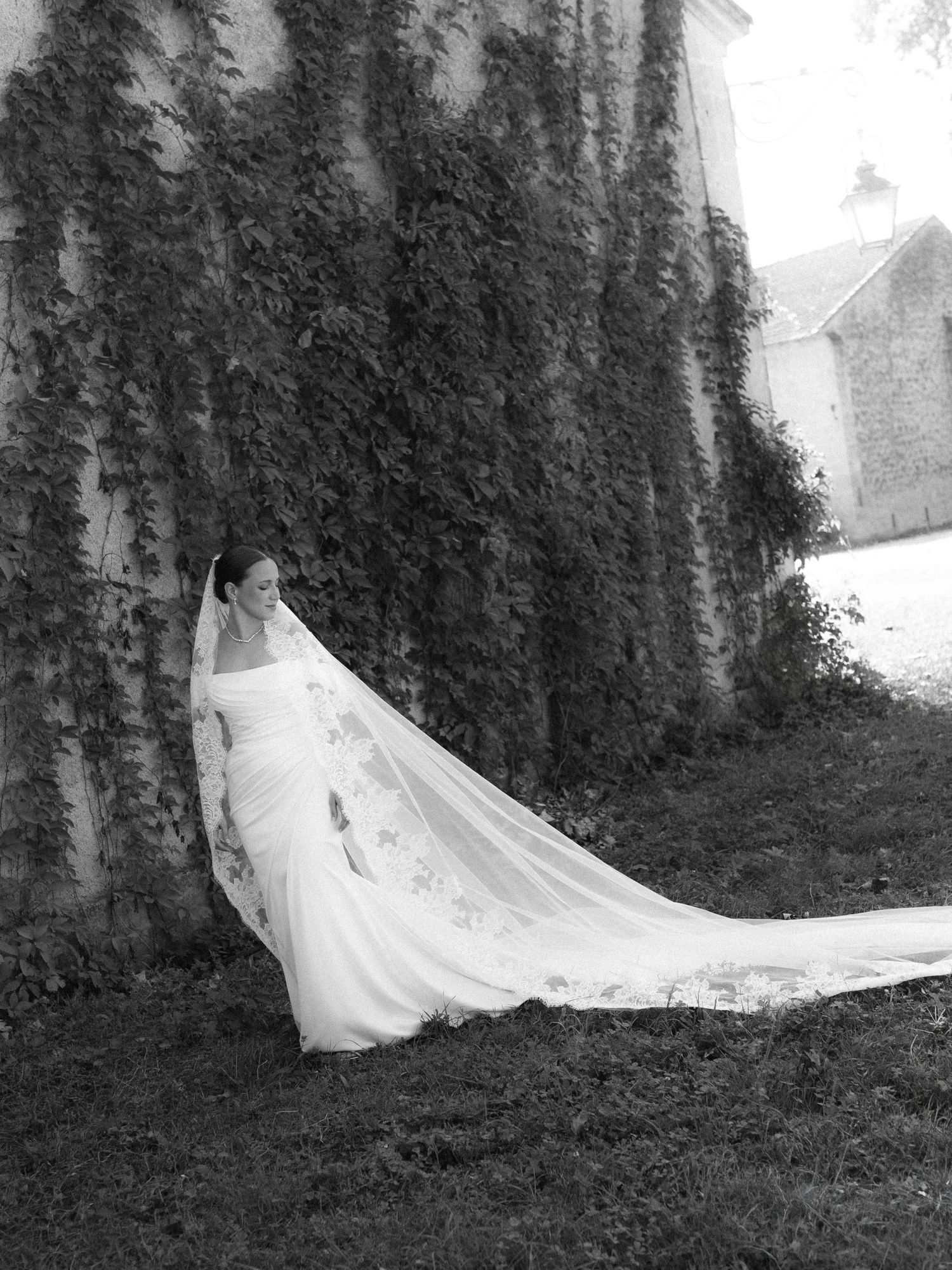 Black and white photo of a bride in a flowing gown and long veil leaning against an ivy-covered stone wall.