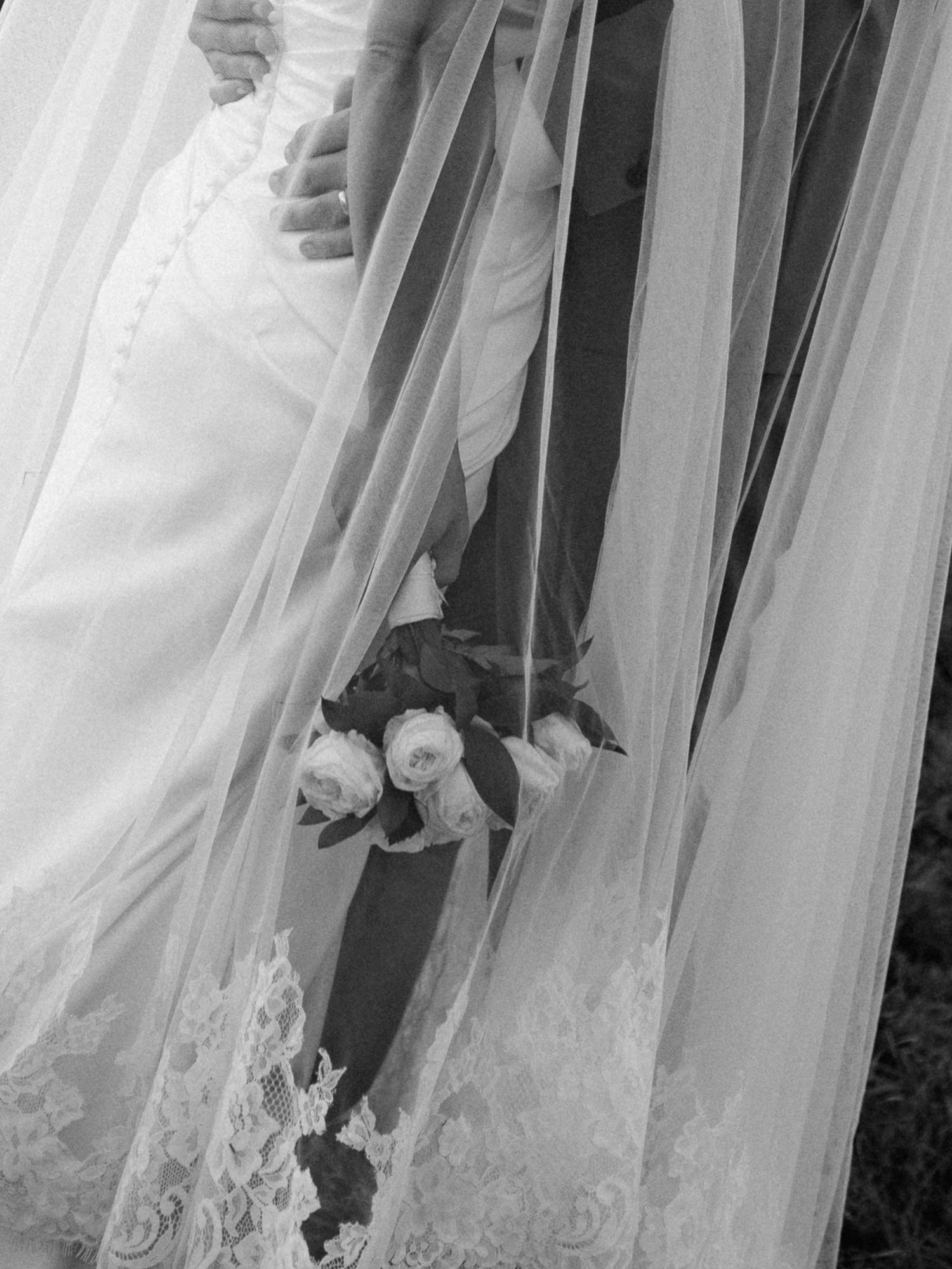 Black and white photo of bride and groom holding a rose bouquet, with flowing wedding veil and lace dress.