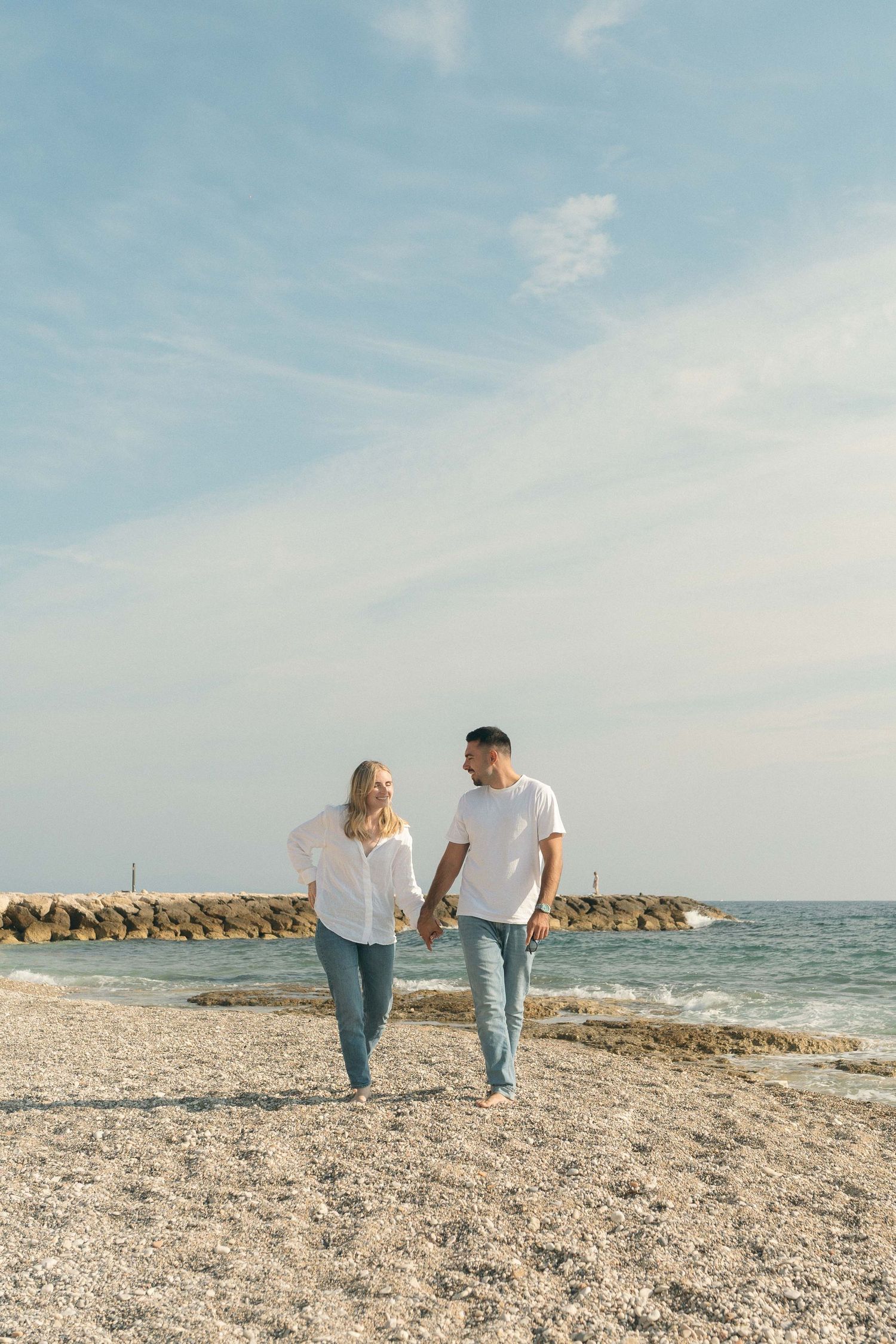 A couple walks hand in hand along a rocky beach shoreline with blue skies and ocean waves in the background.