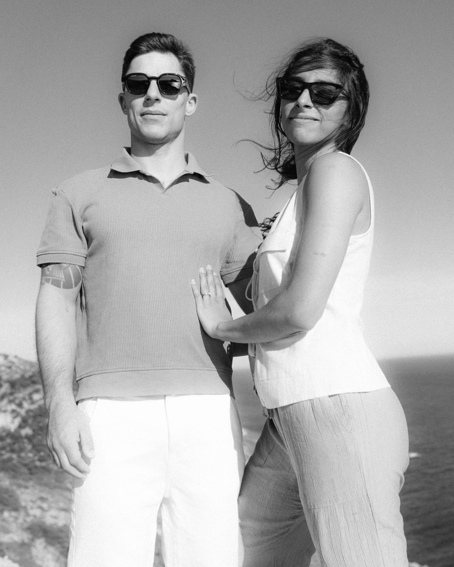 Black and white photo of two people in sunglasses posing together outdoors on a sunny day by the ocean.