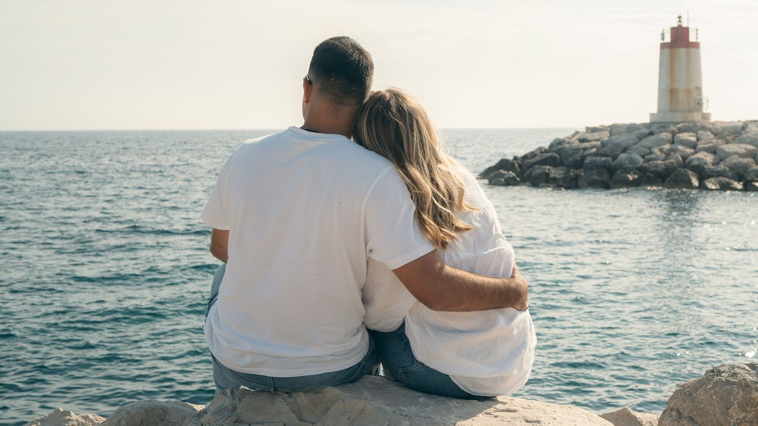 Couple enjoying a romantic sunset moment sitting on a pier by a lighthouse overlooking the Mediterranean Sea.