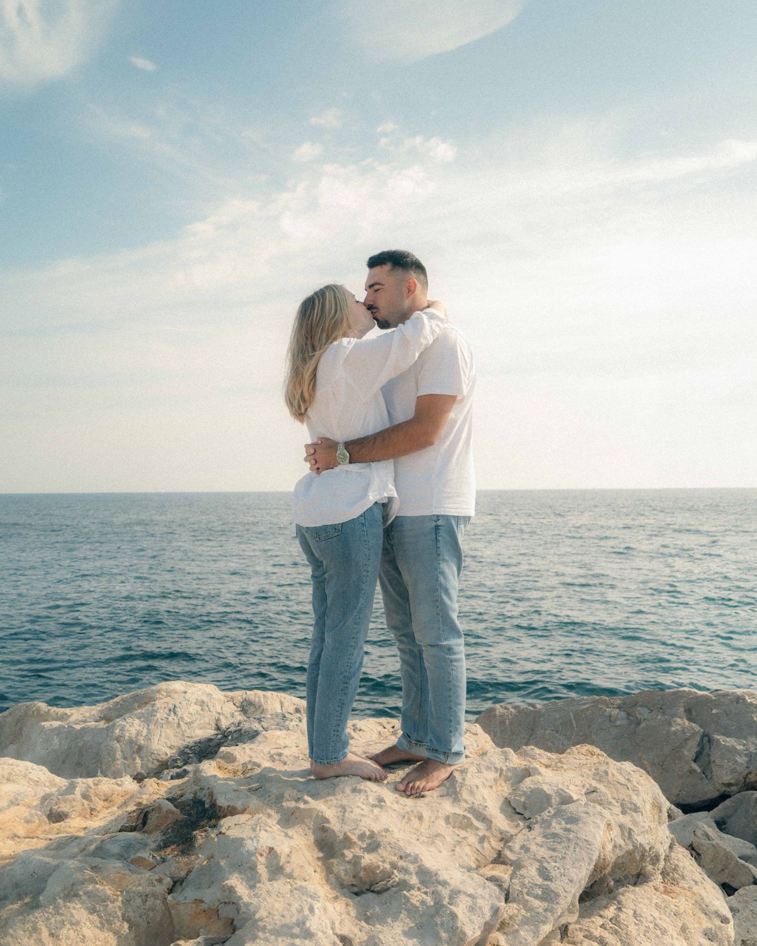 A couple shares a romantic moment together on rocky coastal cliffs overlooking a serene ocean at sunset.
