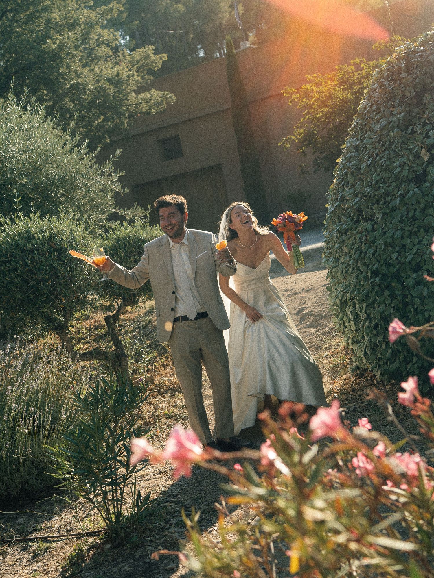 A joyful bride and groom walk through a sunlit garden with lush greenery and colorful flowers on their wedding day.