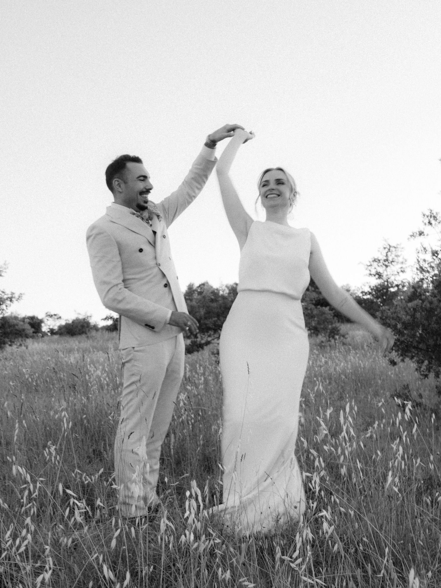 Black and white photo of a couple dancing in a field, the groom spinning the bride in her elegant wedding dress.