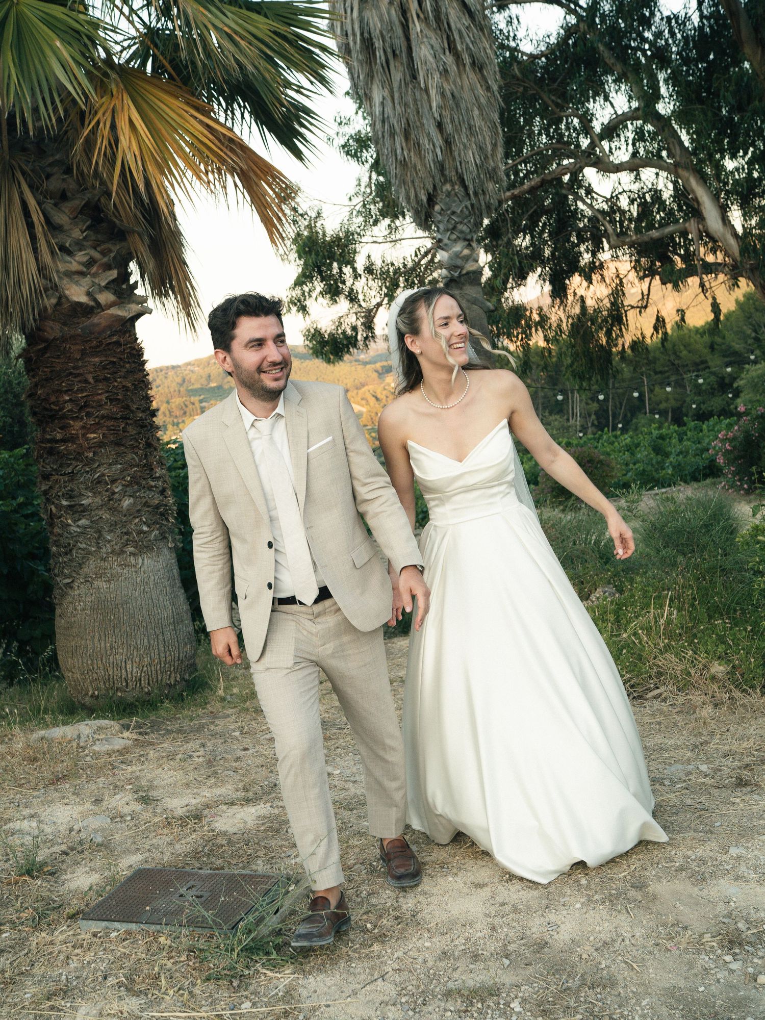 Happy newlyweds walk hand-in-hand outdoors near a palm tree, groom in tan suit, bride in white strapless gown.