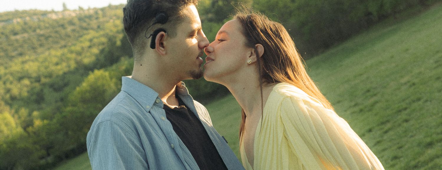 A couple shares a romantic kiss outdoors with lush green hills in the background on a sunny day.