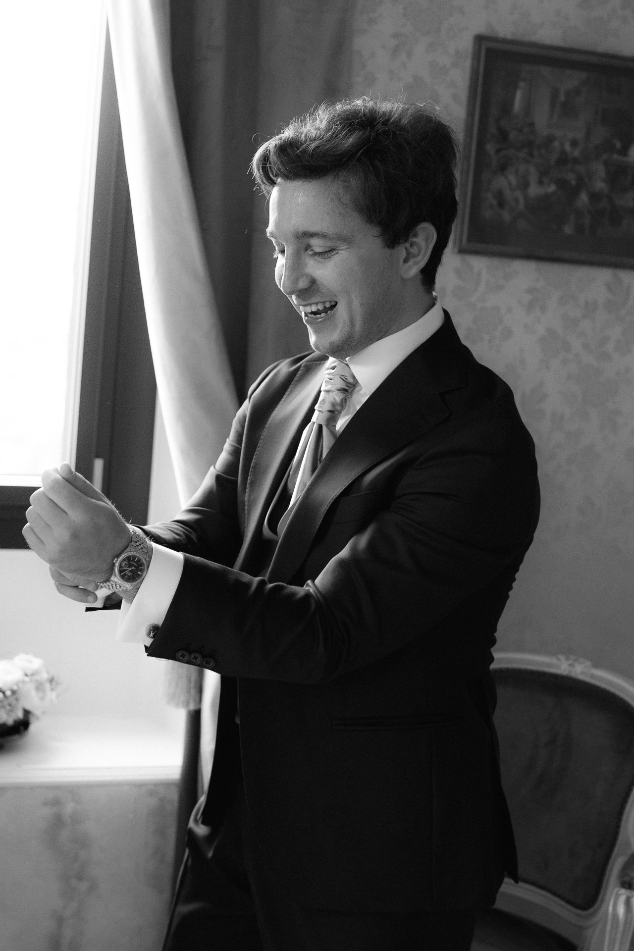 A person in a black suit laughs while sitting in an elegant room with soft lighting.