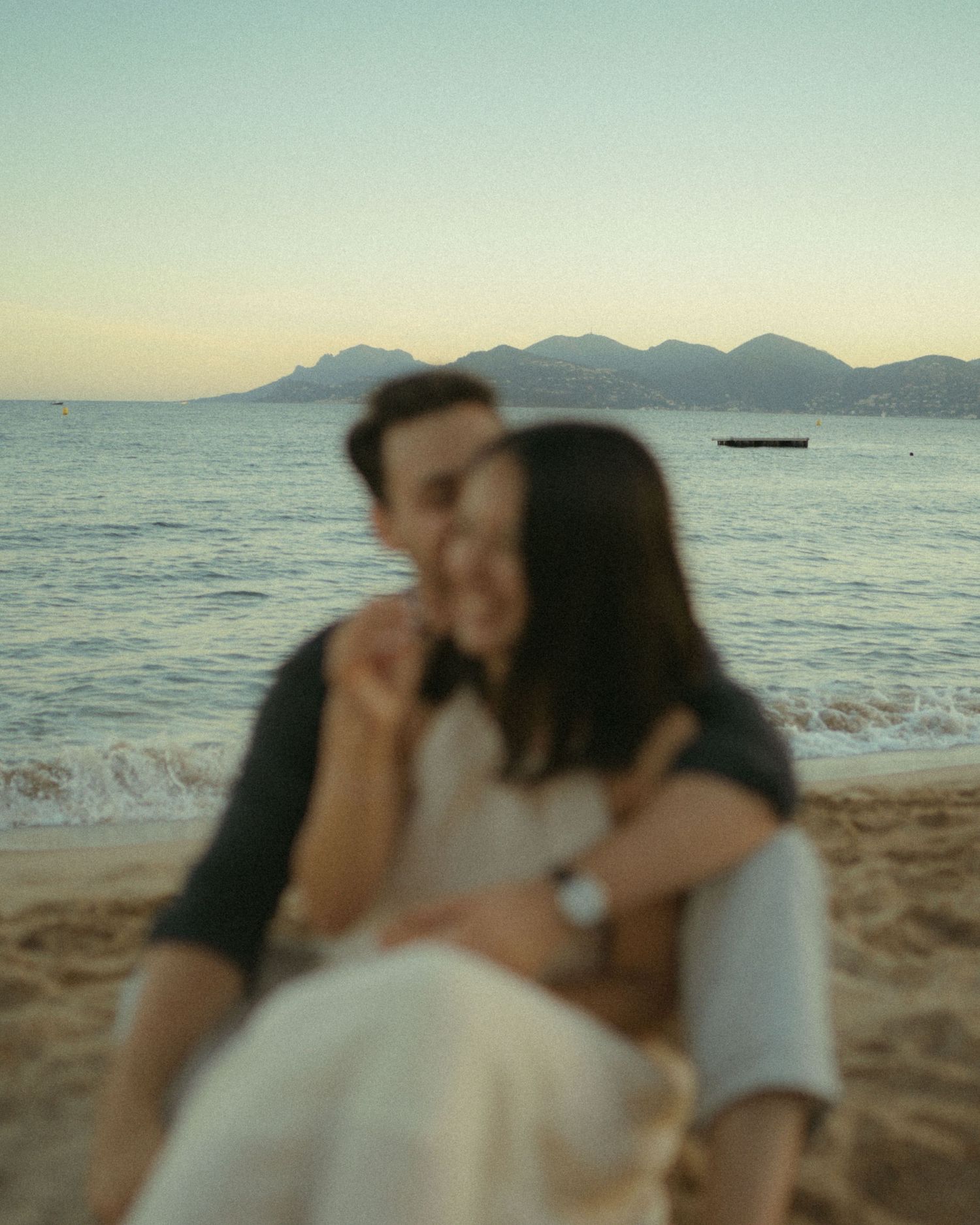 Romantic couple sharing tender moments at sunset on a beach with mountains visible in the distance.