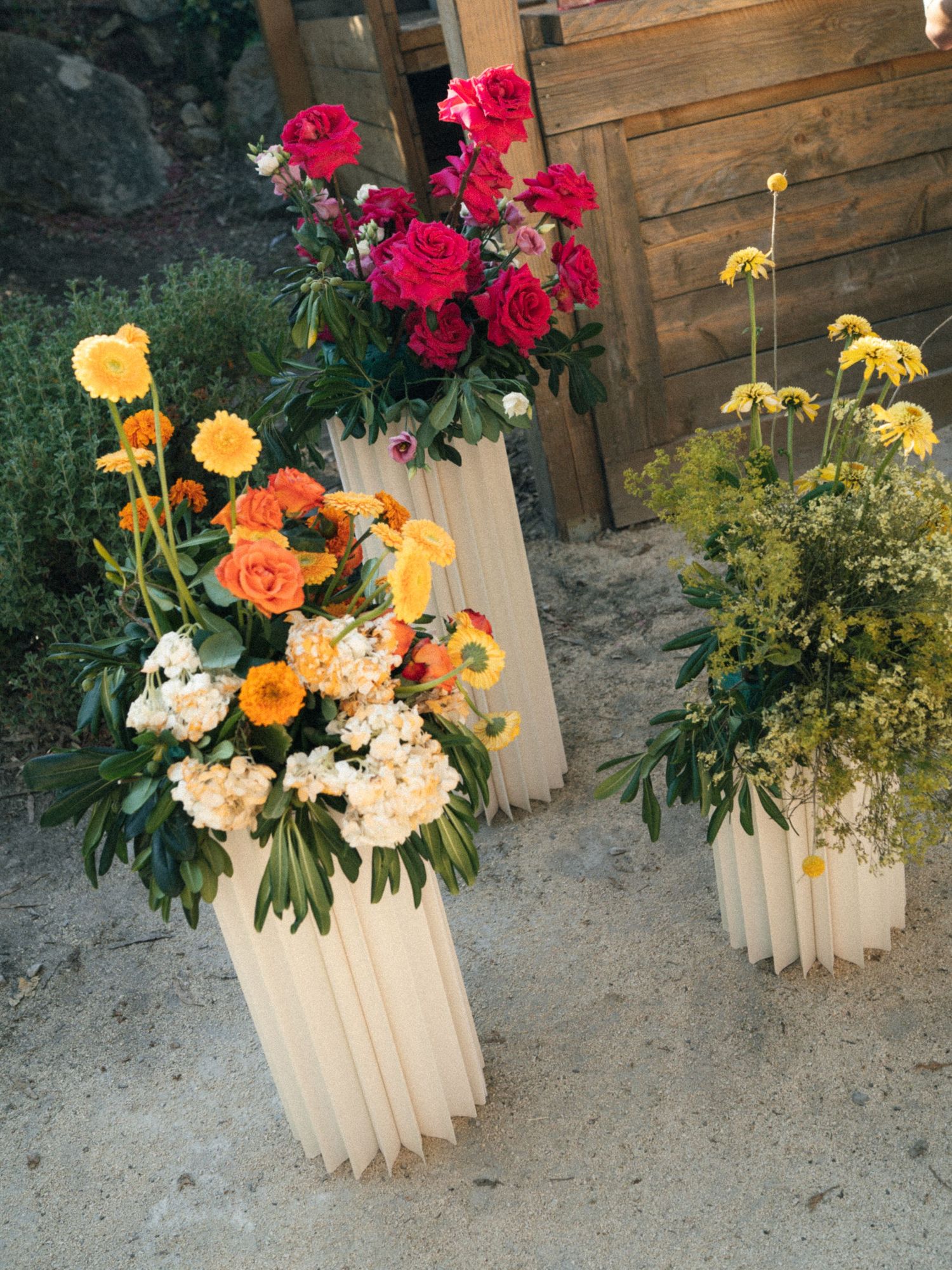 Three tall cream fluted vases with vibrant red, orange, yellow, and white floral arrangements displayed outdoors near a wooden backdrop.