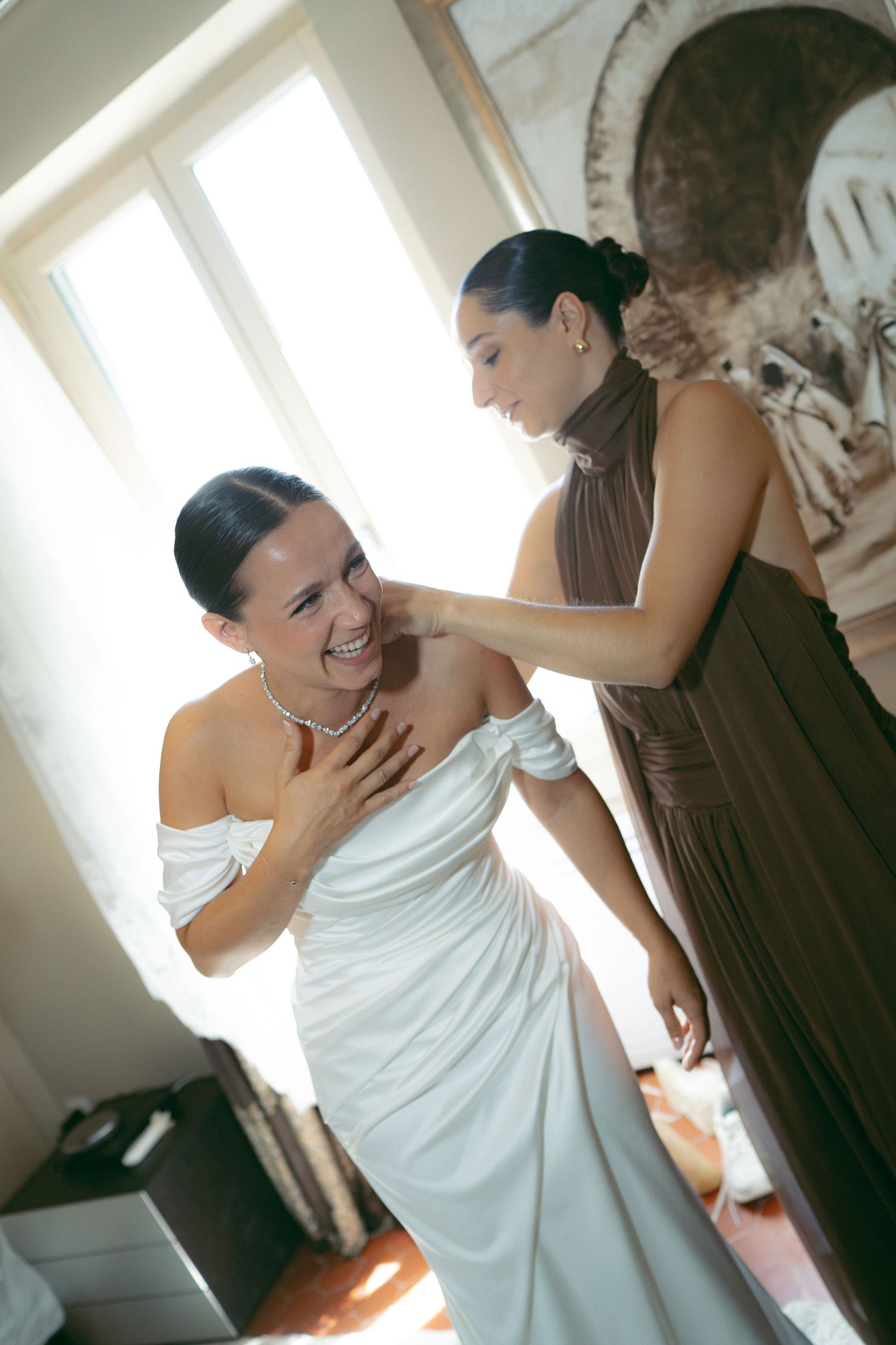 Bride in white off-shoulder gown smiles as bridesmaid in brown dress helps her prepare for wedding ceremony.
