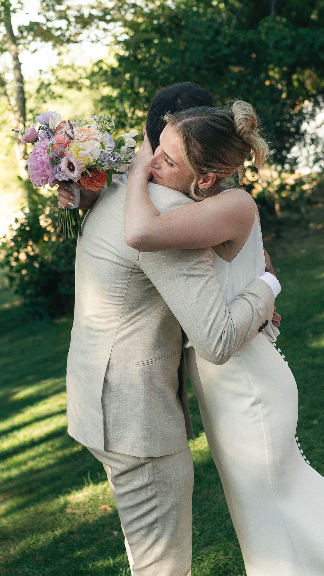 A bride and groom share an emotional embrace outdoors, she holds a colorful bouquet, both wearing elegant cream attire.