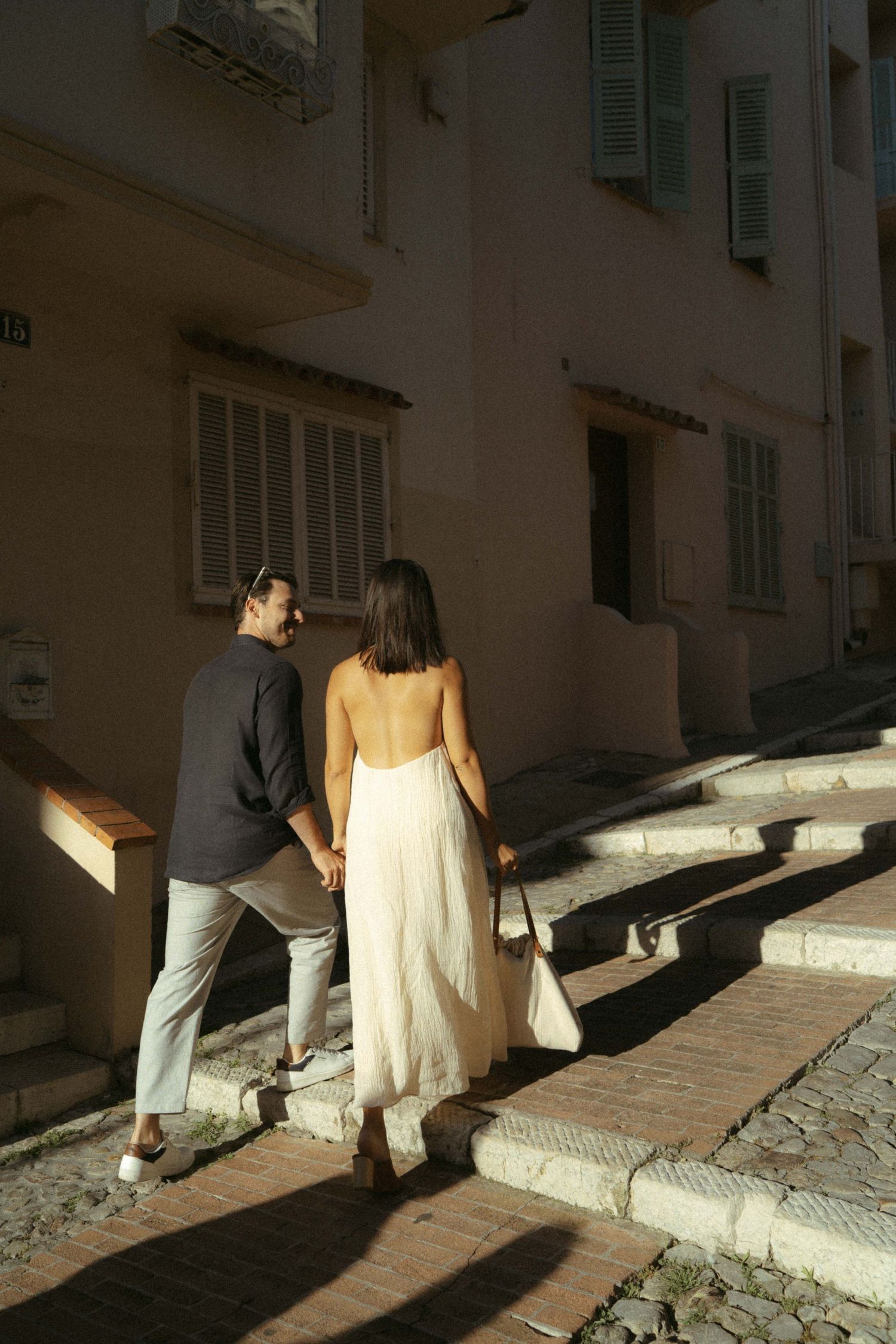 A couple walks hand in hand up a sunny Mediterranean street with white walls and green shutters.