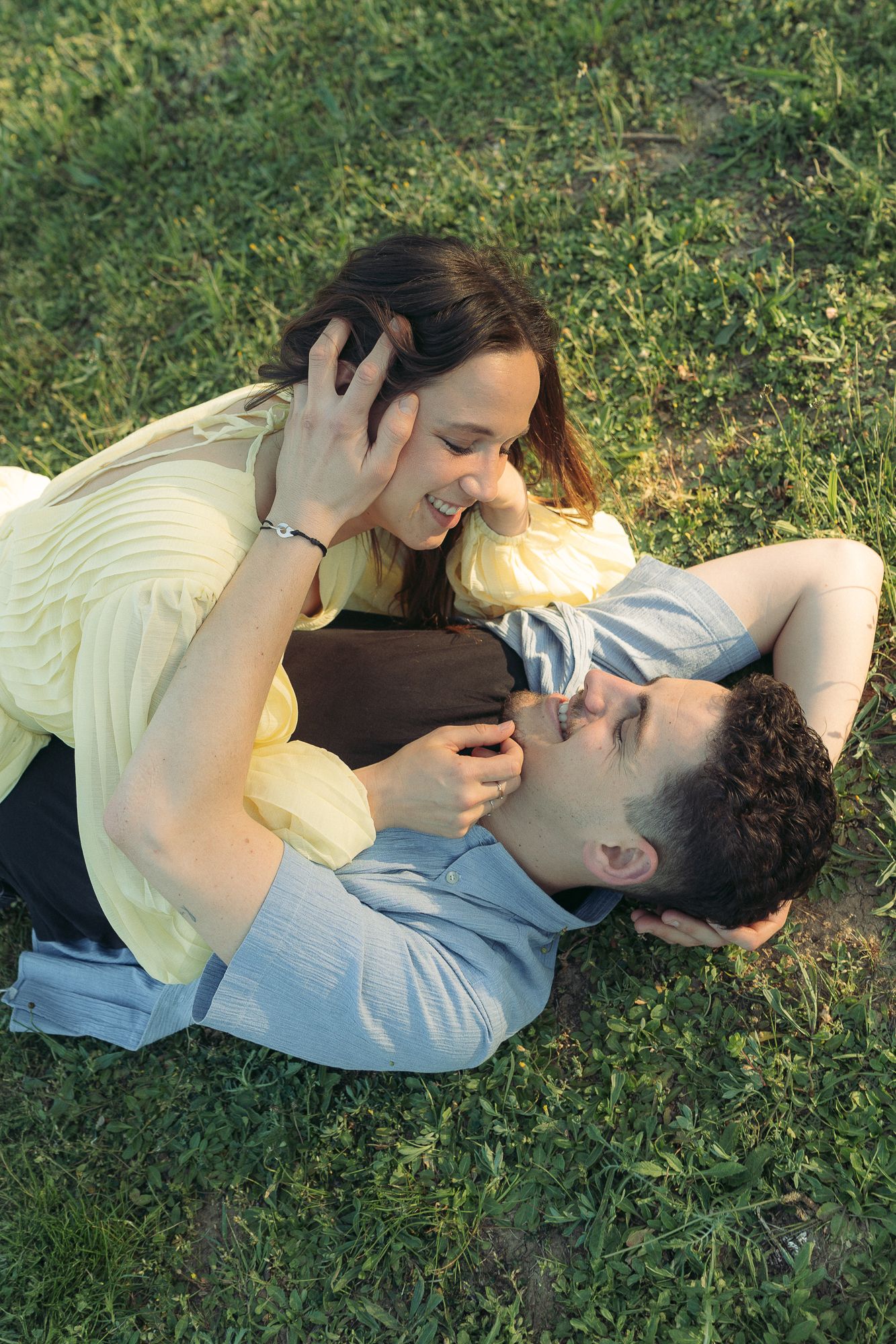 A happy couple relaxing on green grass, with the man lying down and the woman leaning over him, smiling intimately.