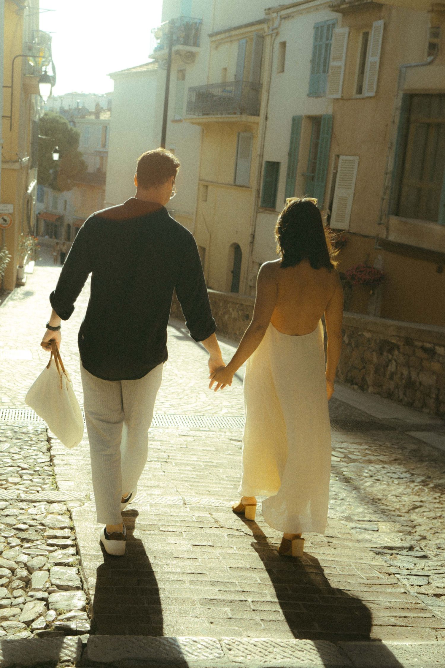 People walking on a cobblestone street cast long shadows in warm evening sunlight.
