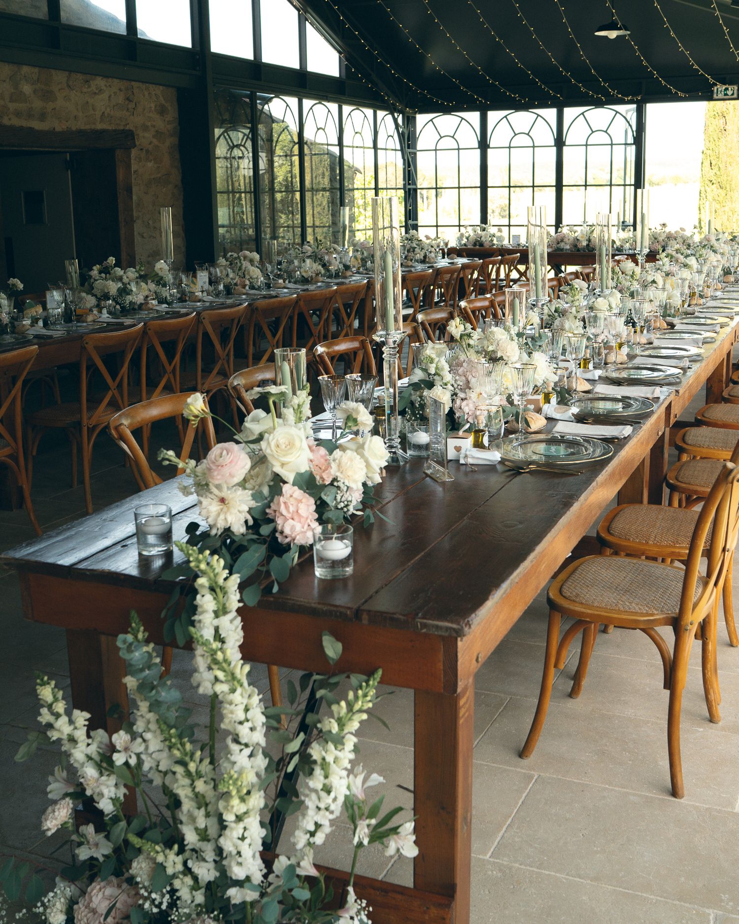 Elegant wedding reception venue with long wooden tables adorned with white floral centerpieces and crossback chairs.