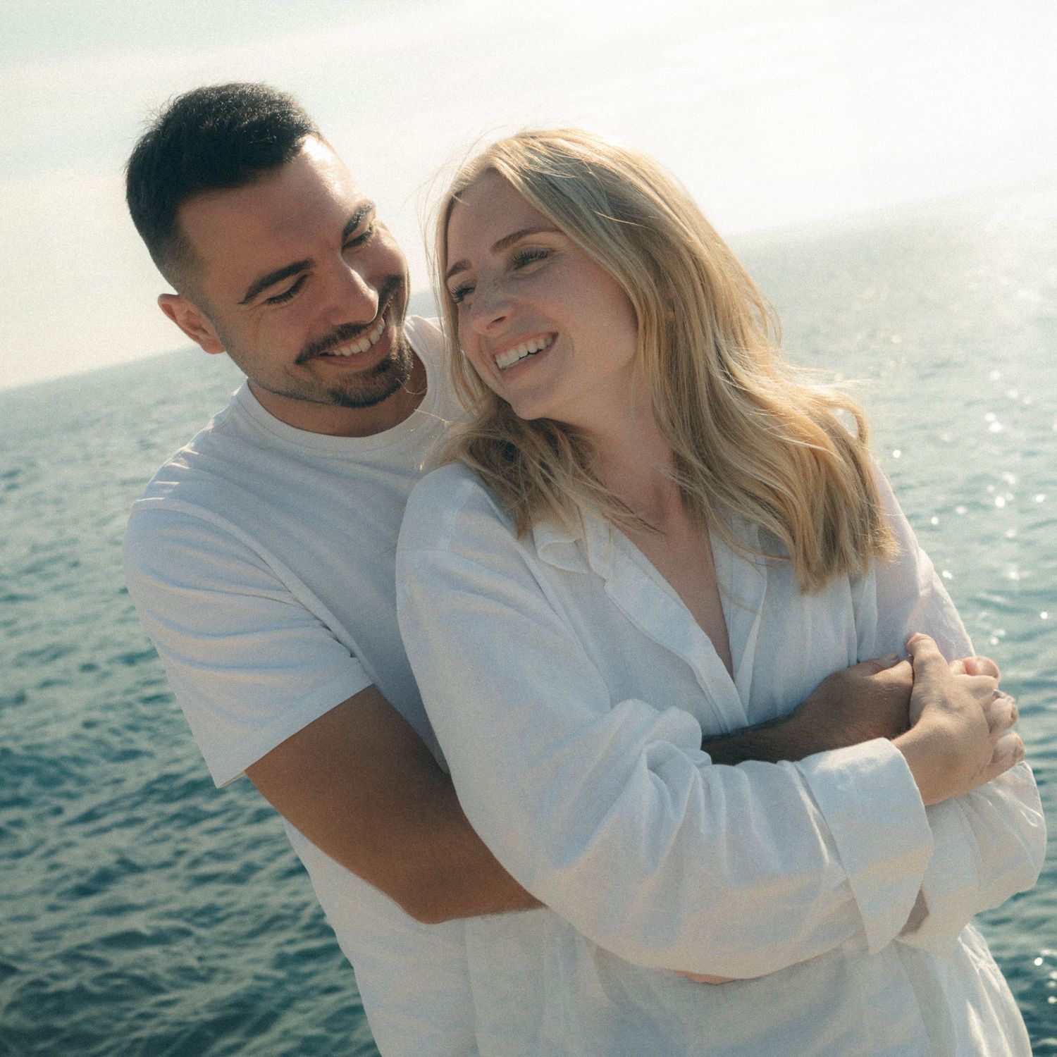 Couple in white clothing embracing and laughing together by the ocean during golden hour.