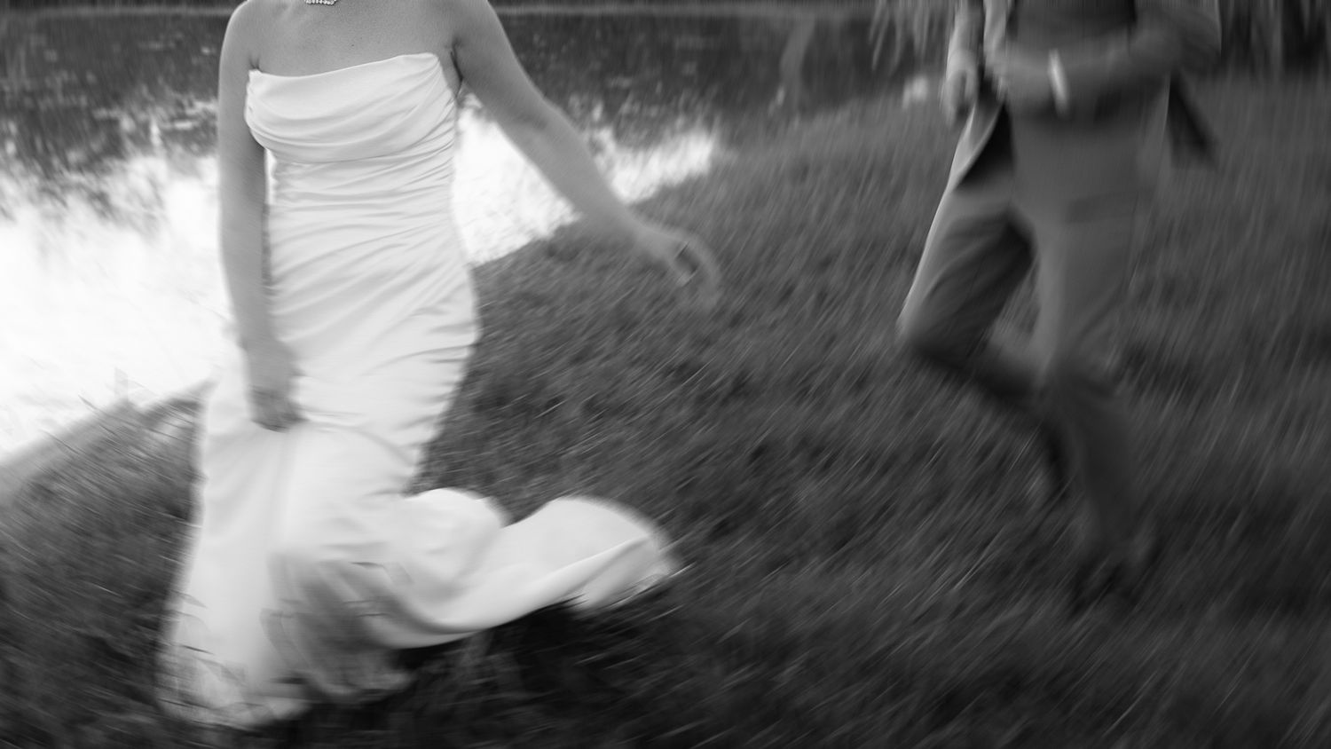 Black and white photo of a bride in white gown running outdoors near water with another person.