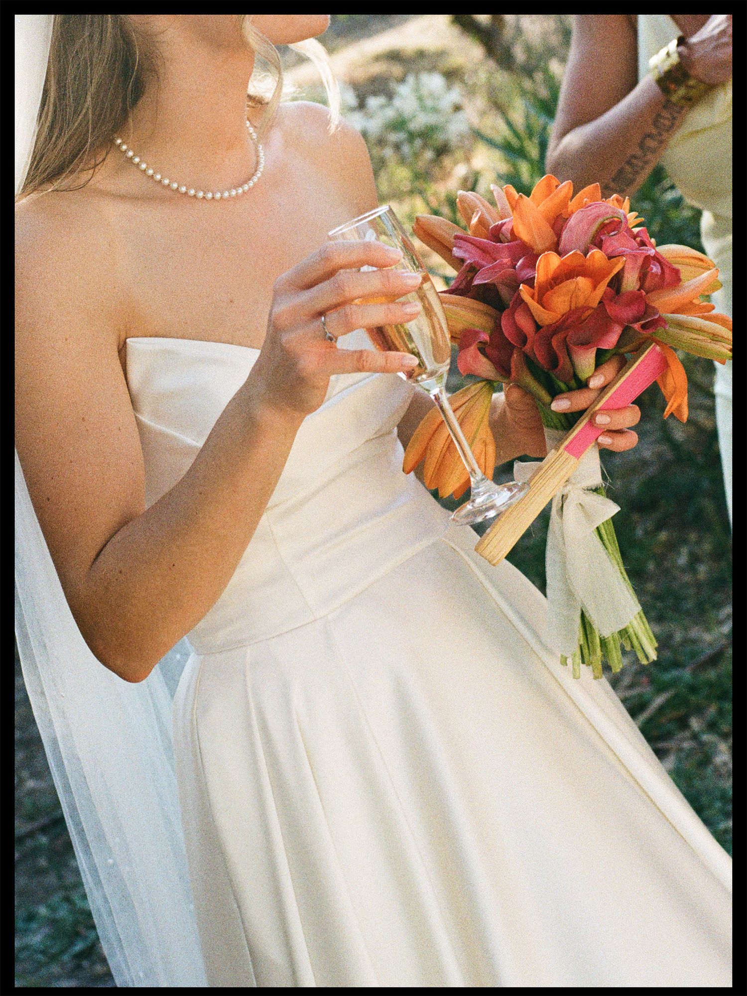 Bride in strapless white gown holding champagne flute and vibrant orange tropical bouquet with pearl necklace.