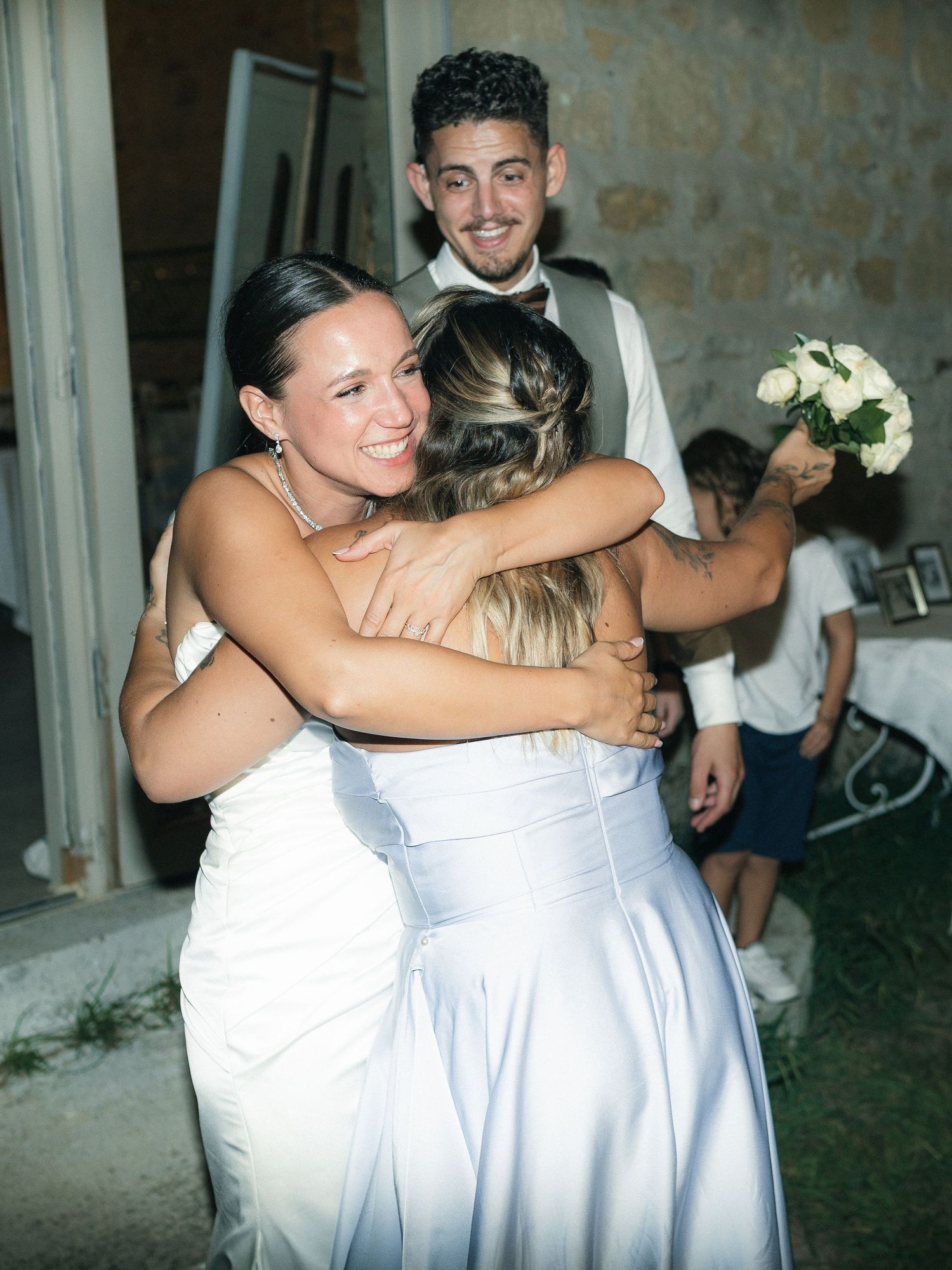 Bride in white dress hugging guest in blue gown at outdoor wedding reception while groom smiles behind them.