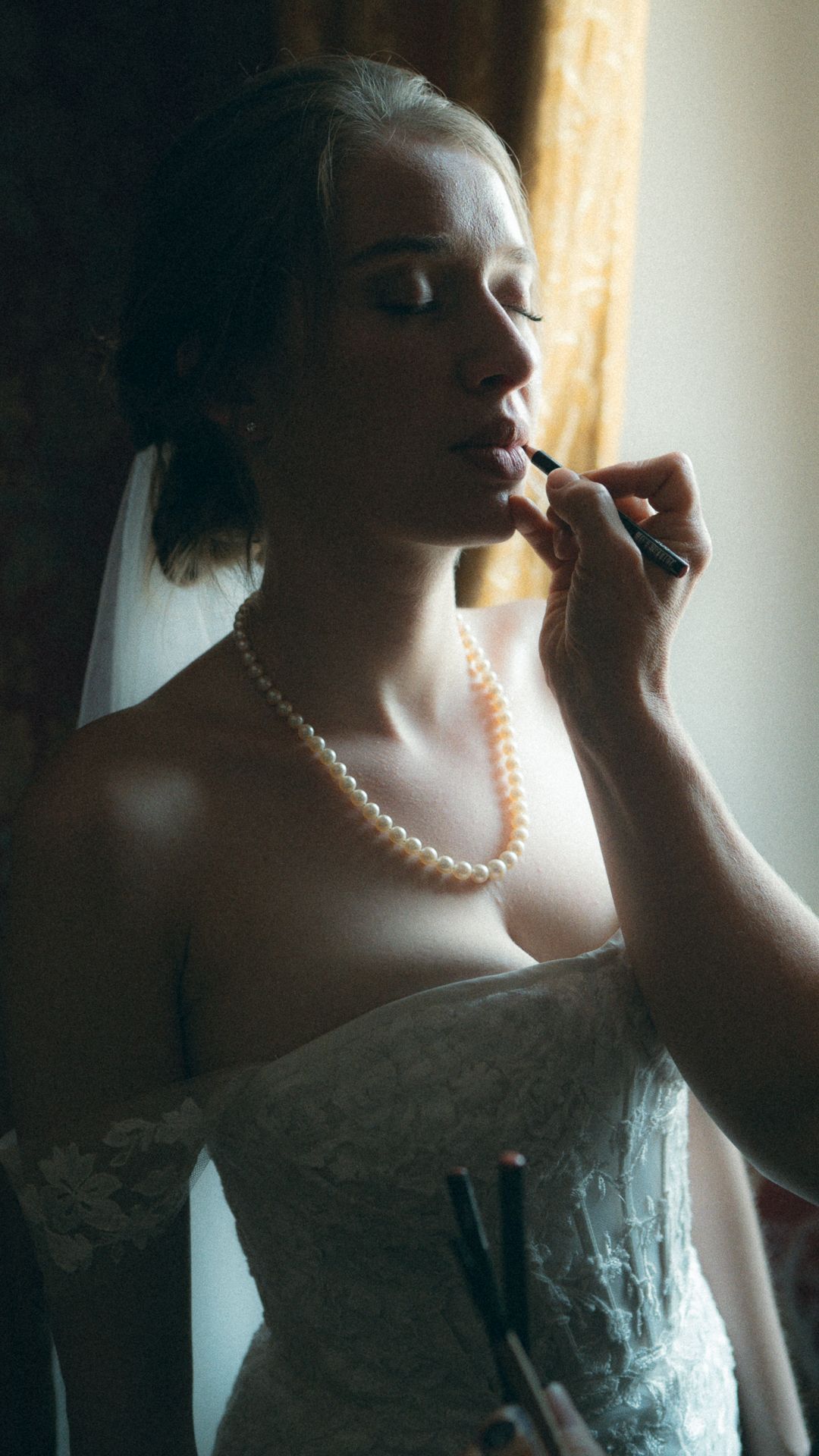 A bride in a white lace dress and pearl necklace has makeup applied during wedding preparations.