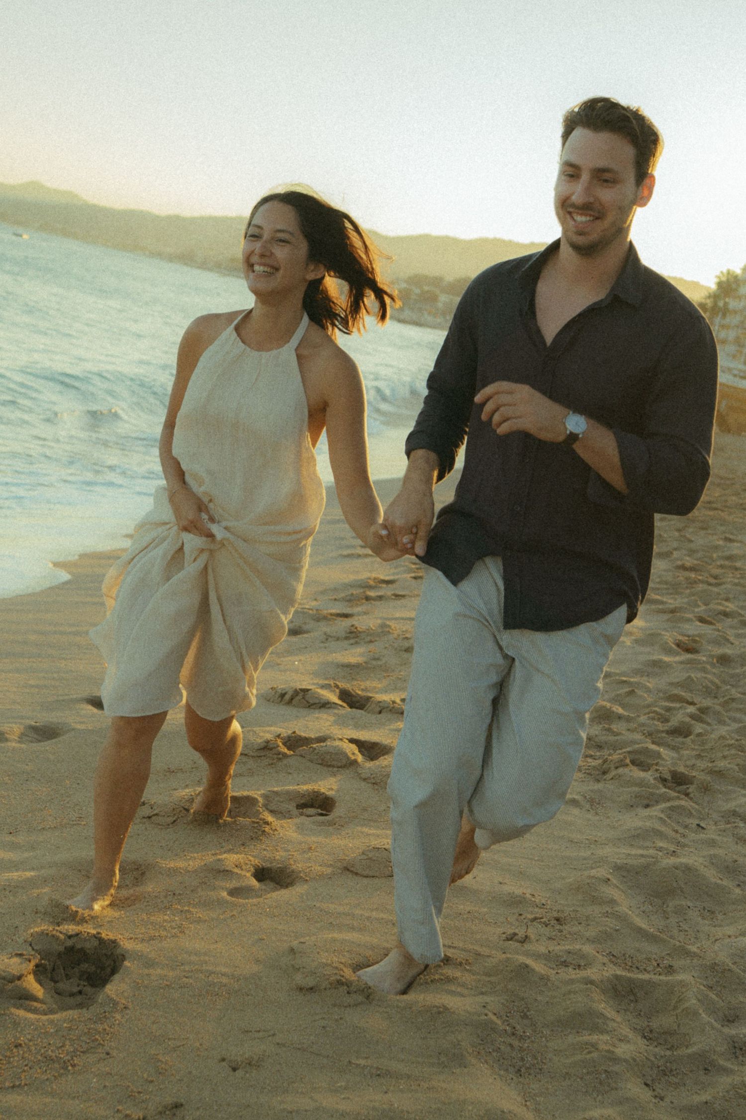 A couple playfully runs and laughs together along a sandy beach at sunset, creating joyful moments by the ocean.