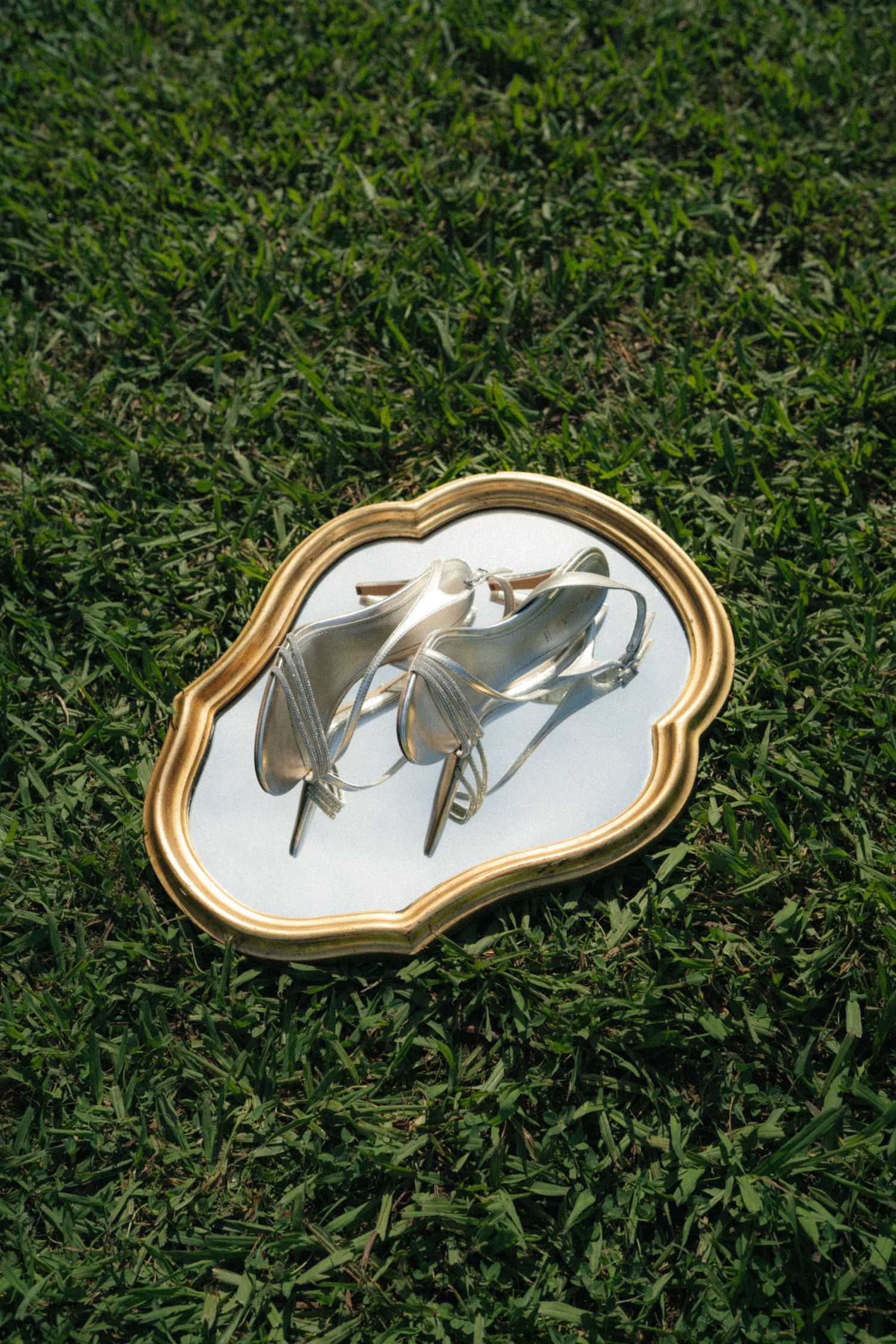 Gold-framed ornate mirror lying on green grass reflecting silver strappy heeled sandals.