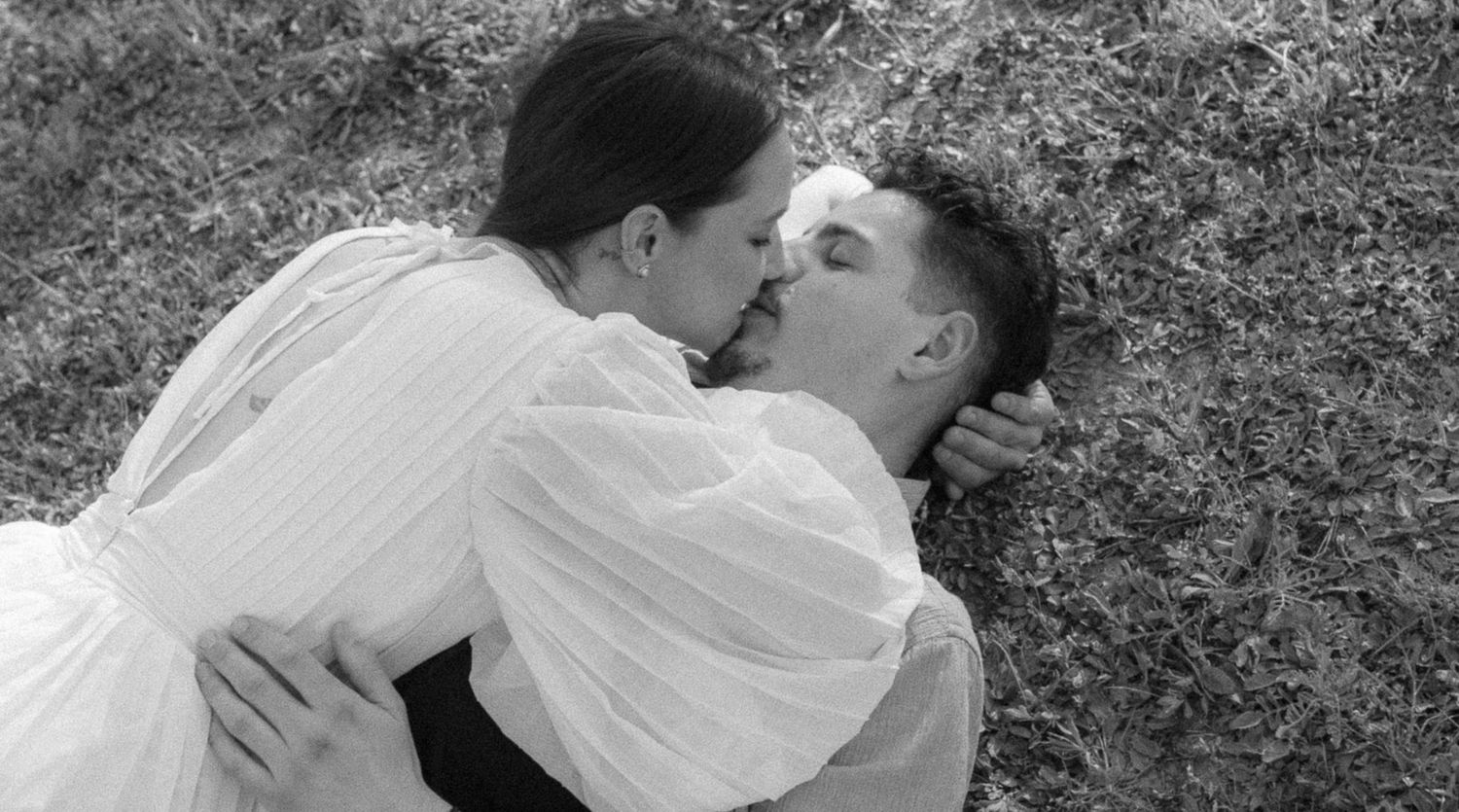 Black and white photo of a couple kissing while lying on grass, dressed in white clothing.